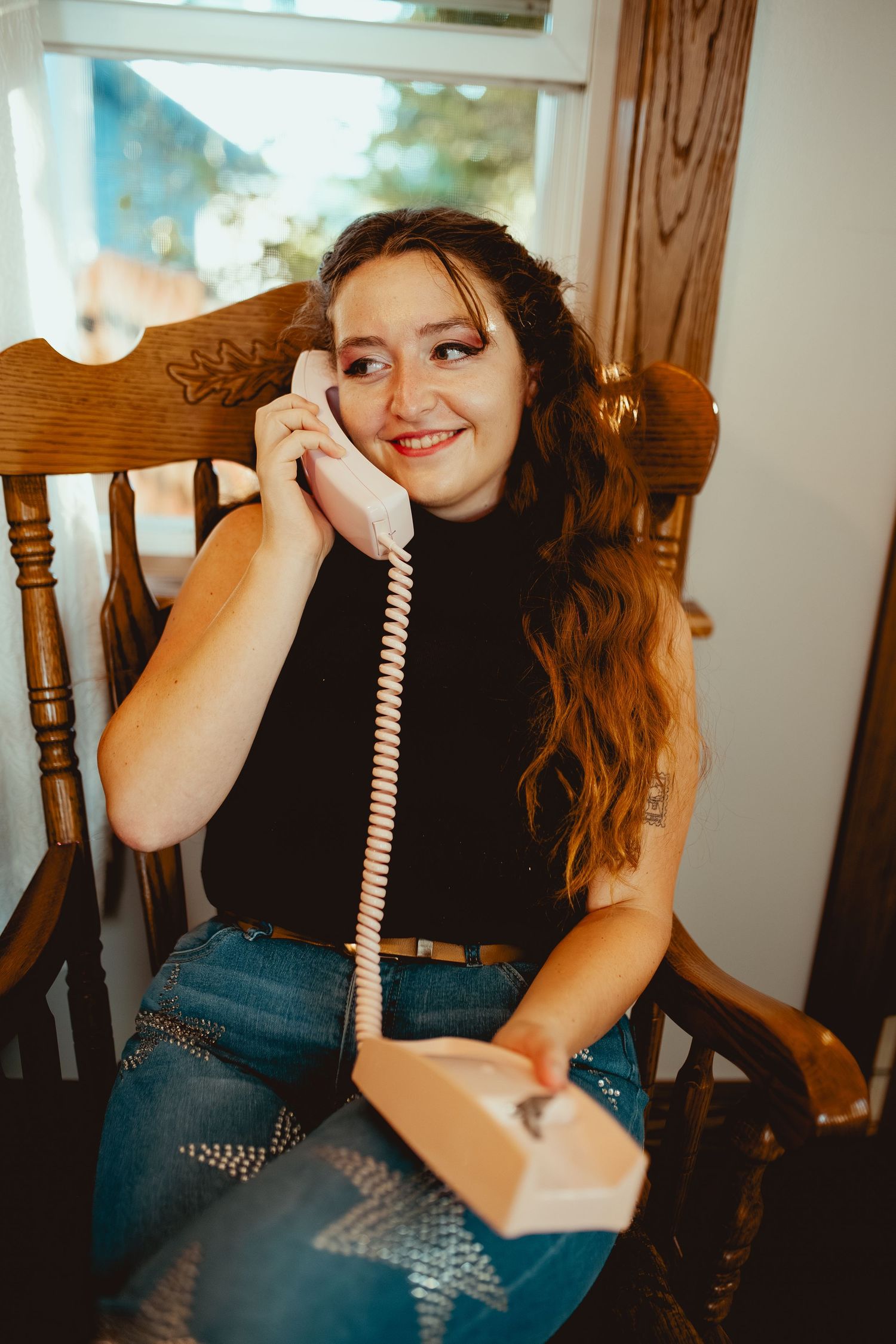 Person in black top and denim sitting on wooden chair holding corded telephone receiver in casual indoor setting.