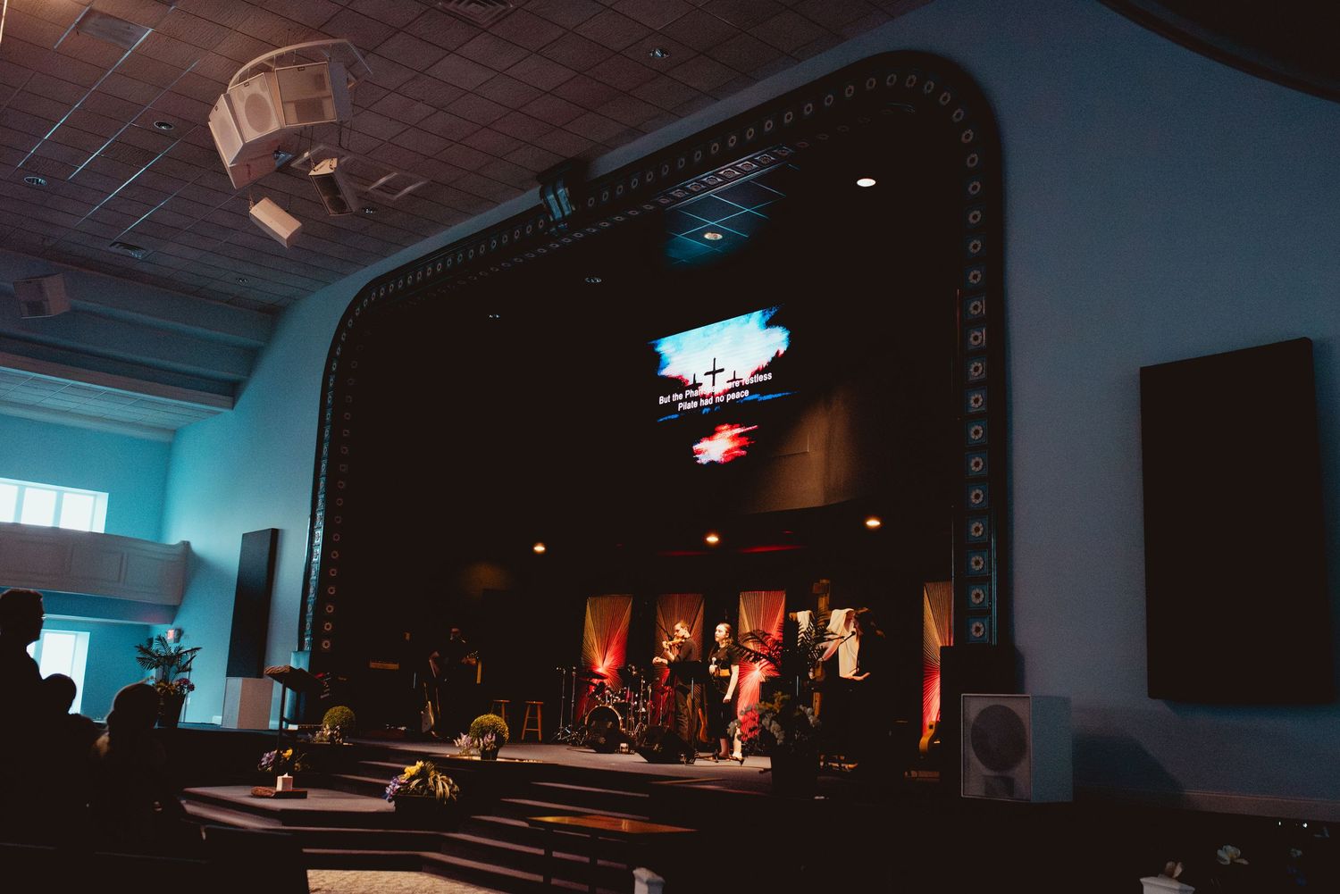 A dimly lit stage with musicians performing in a church or auditorium setting with blue ambient lighting.