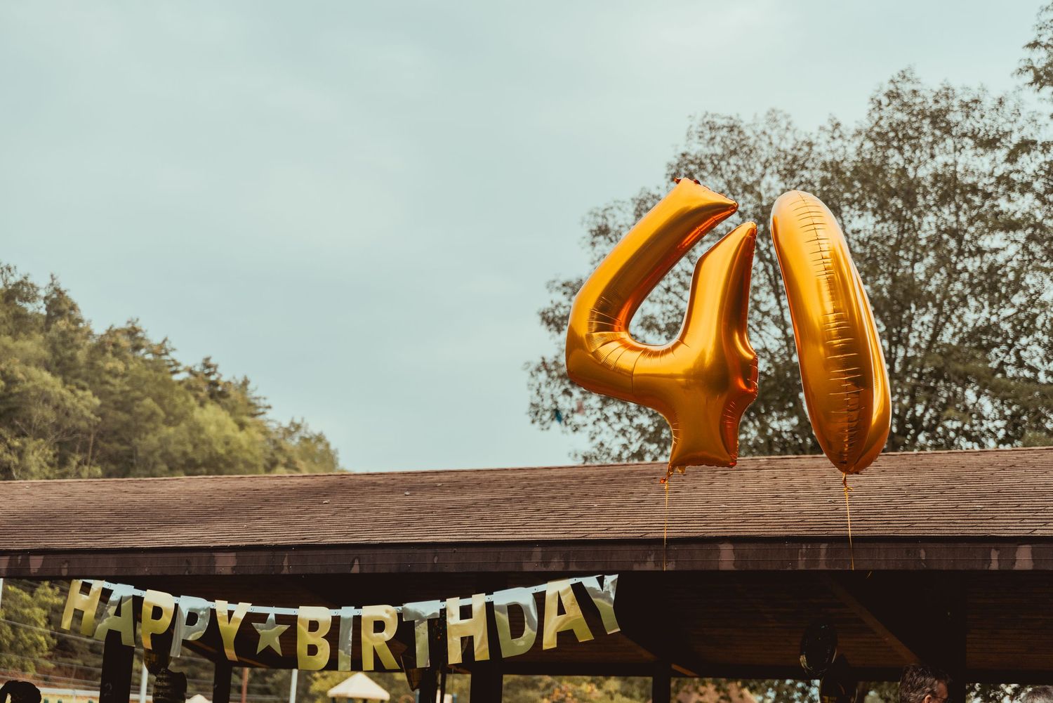 Colorful birthday party decorations including orange balloons and a 'Happy Birthday' banner hang from a wooden structure outdoors.