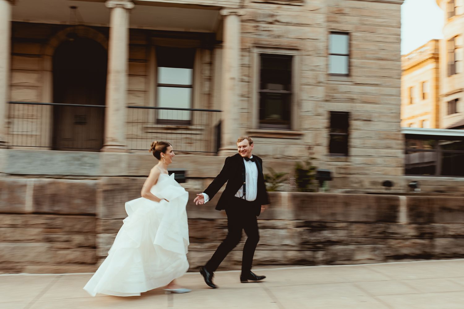 A bride in a flowing white gown walks alongside someone in formal attire near a stone building.