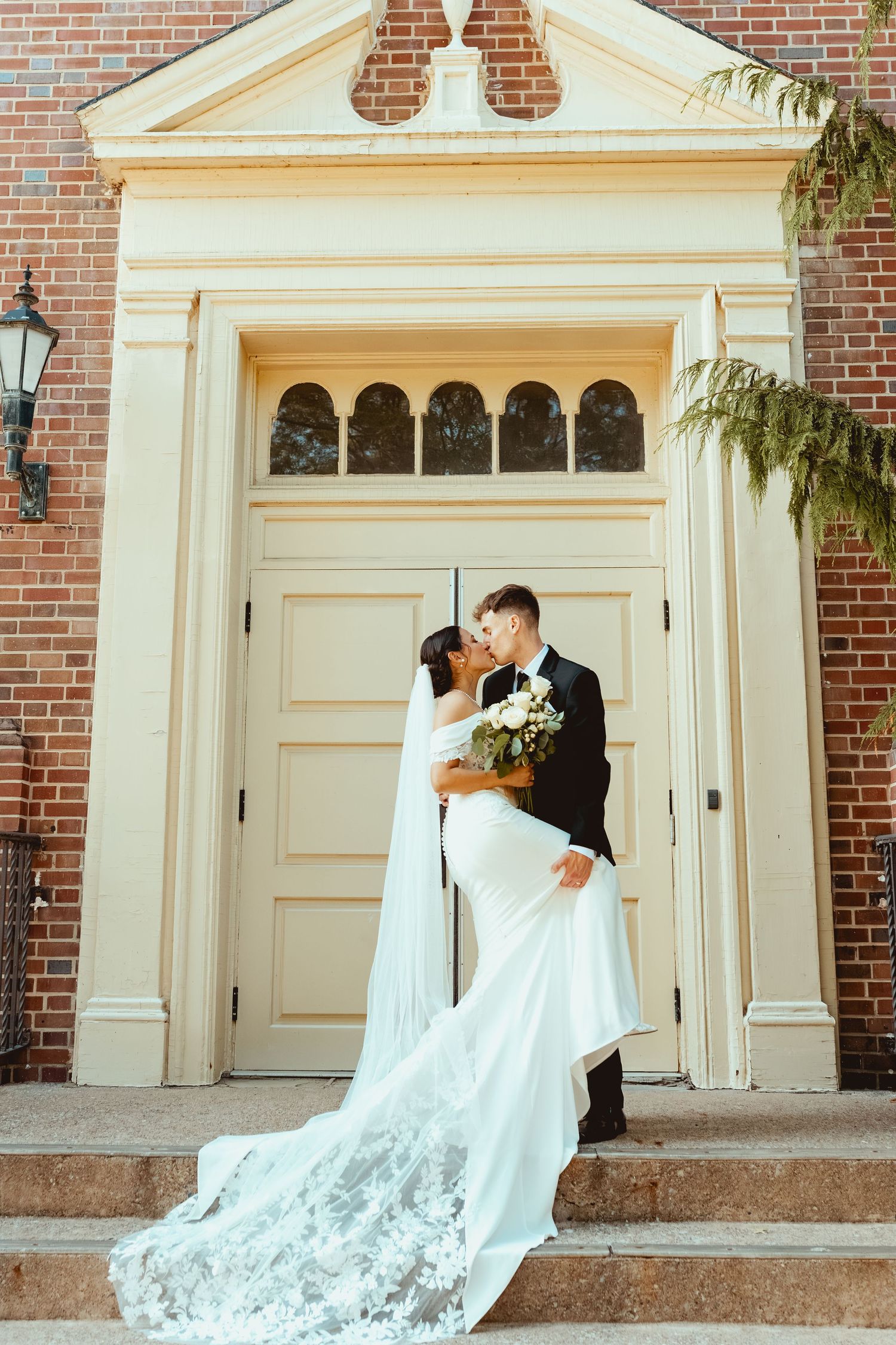 A romantic wedding couple embraces in front of a classic brick church with elegant white doors and columns.