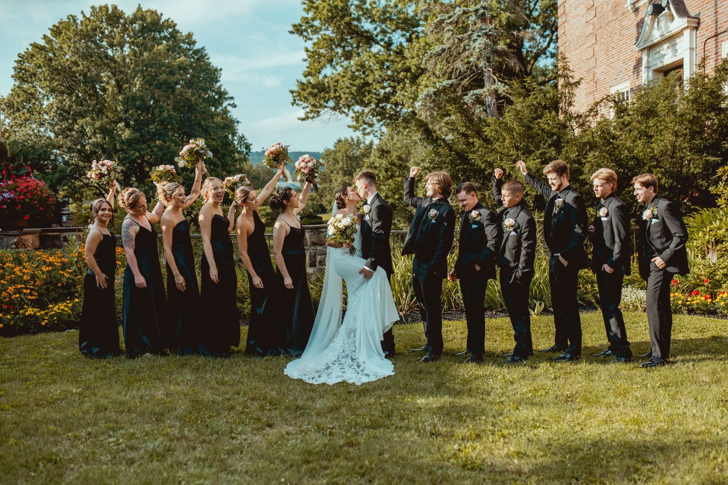 Wedding party stands in formation on grass with trees and buildings in background during summer day.