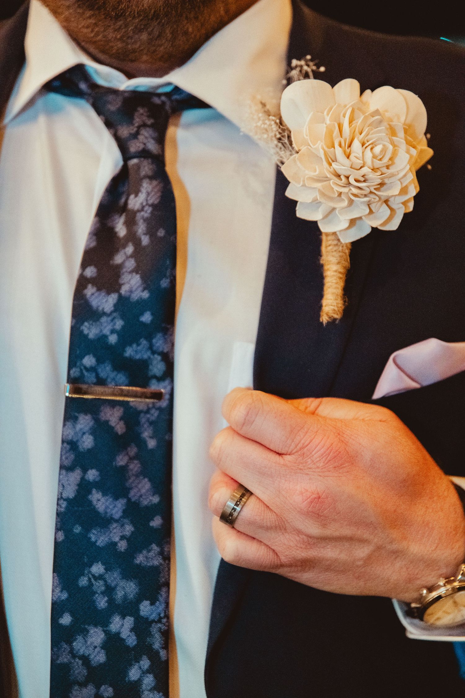 Close-up of groom in navy suit with floral tie, boutonniere, and wedding ring on his hand.