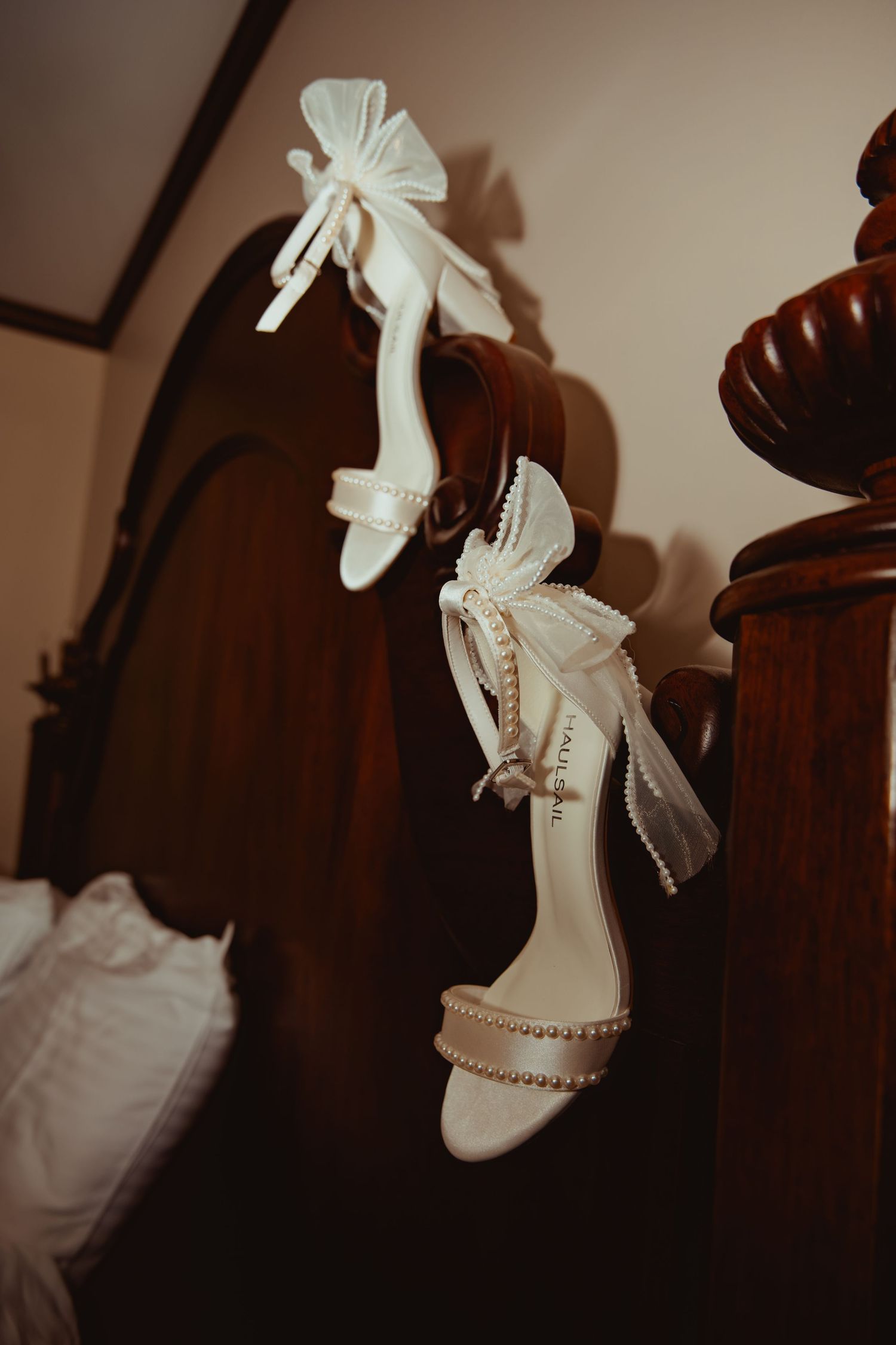 Elegant white bridal heels with pearl embellishments and bow details hanging on a wooden headboard.