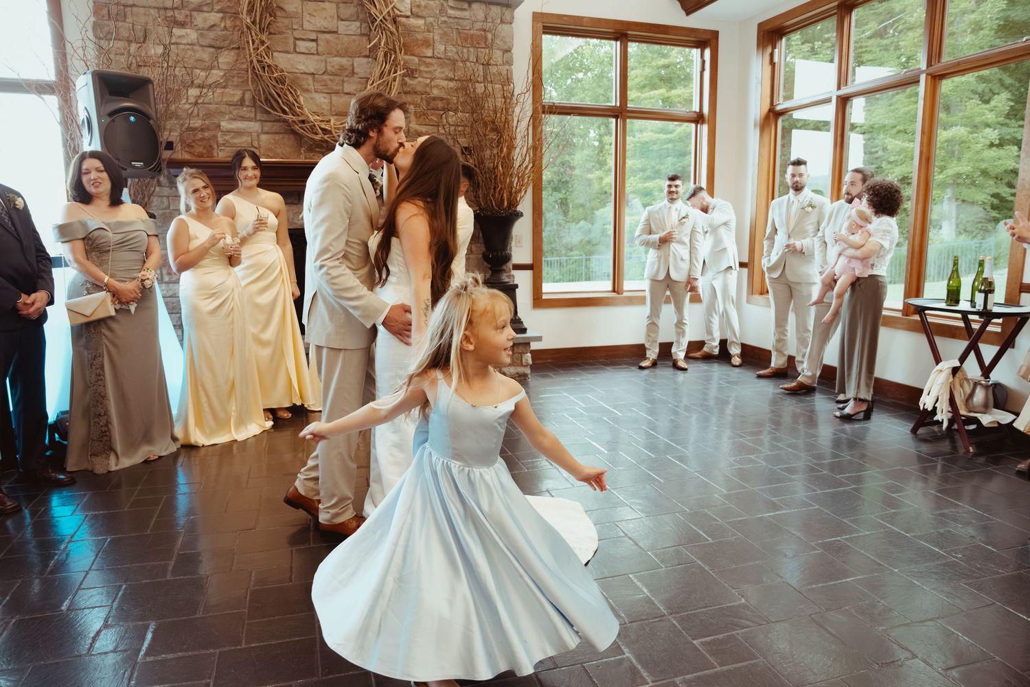 Flower girl twirls in blue dress as newlyweds share first dance surrounded by wedding party.
