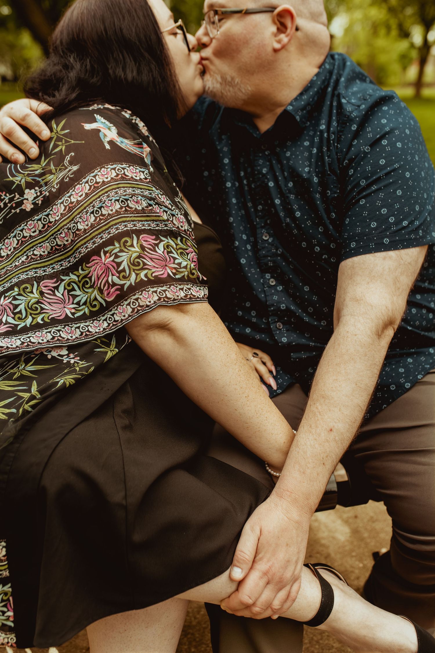 Couple sharing a tender kiss while holding hands outdoors in a park setting.