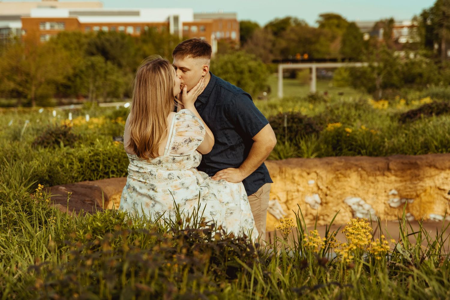 A couple shares a tender kiss while sitting on a stone bench surrounded by lush greenery during golden hour.