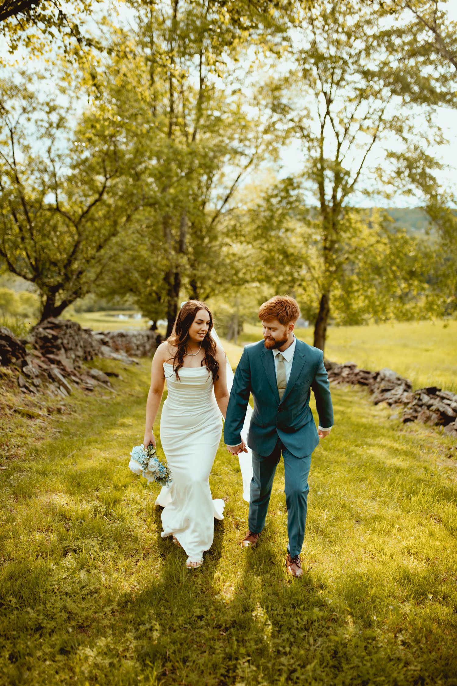 A couple in wedding attire walks together on green grass with a rustic stone wall and trees behind them.