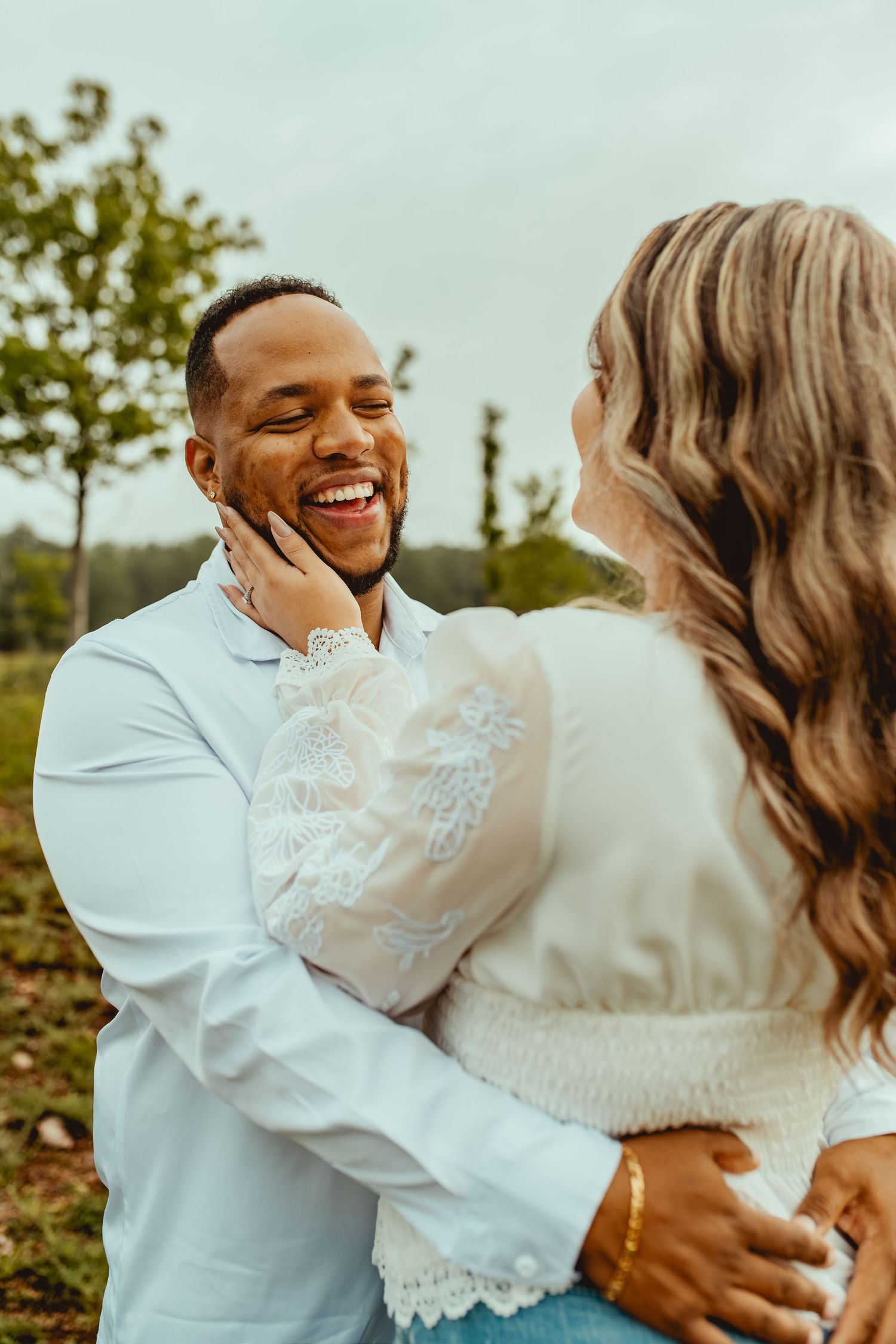 A joyful couple embraces outdoors, the woman tenderly touching the laughing mans face in a romantic moment.