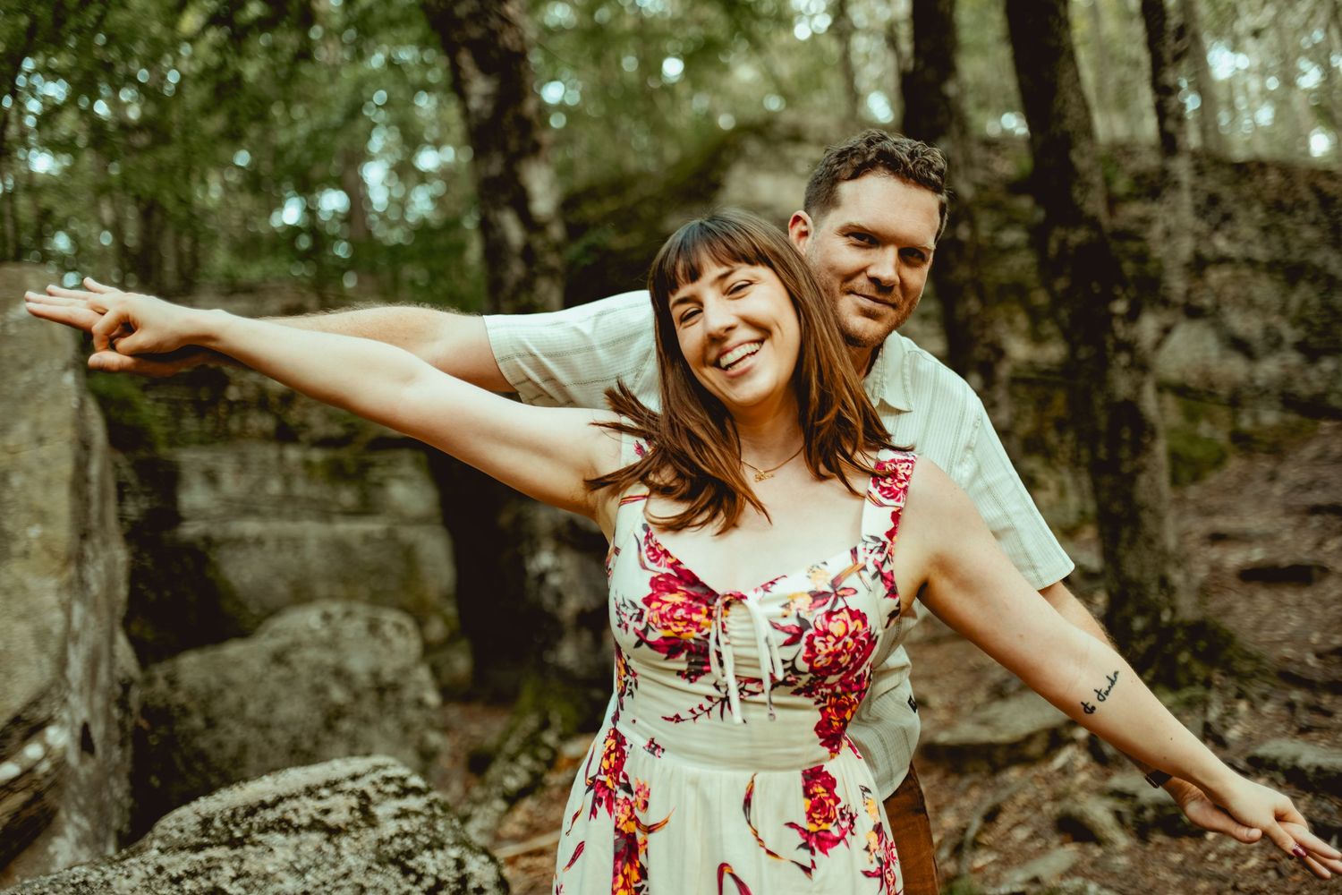 Joyful couple posing in a forest, woman in floral dress with arms outstretched, man smiling behind her near rocky ruins.