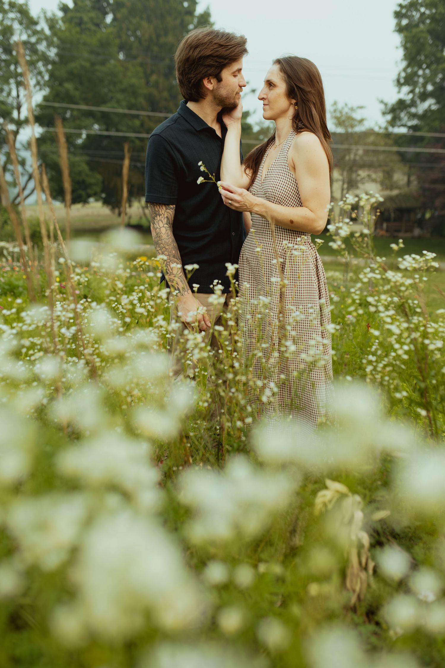 Couple standing in a field of white wildflowers, gazing at each other romantically, man with tattoos in black shirt.