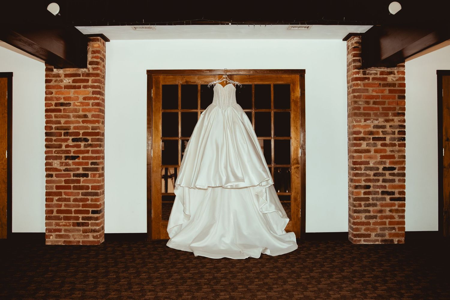 Elegant white ball gown wedding dress hanging on wooden doors between brick pillars in a rustic venue.
