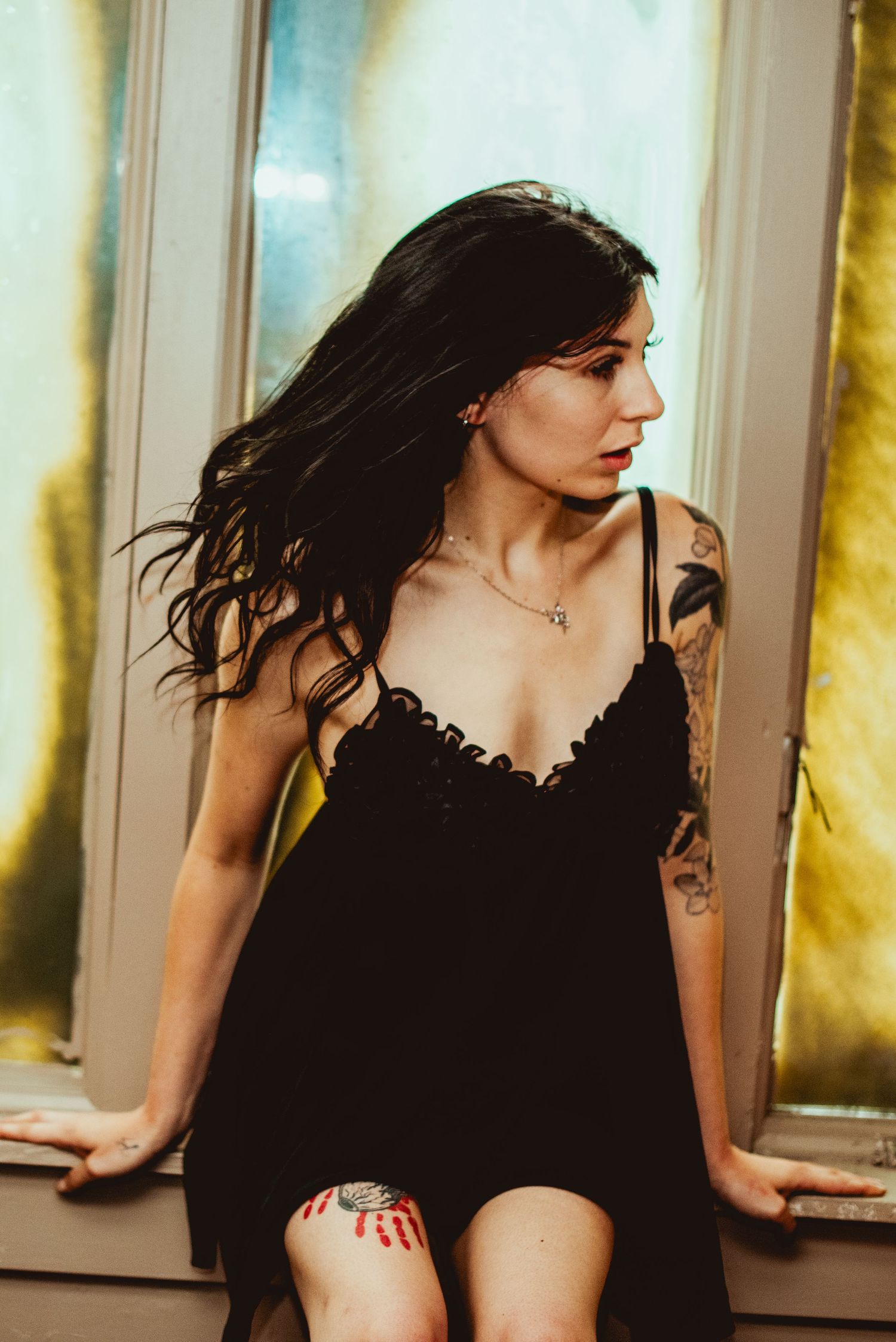 Woman in black dress poses near window with dramatic lighting and flowing dark hair in motion.