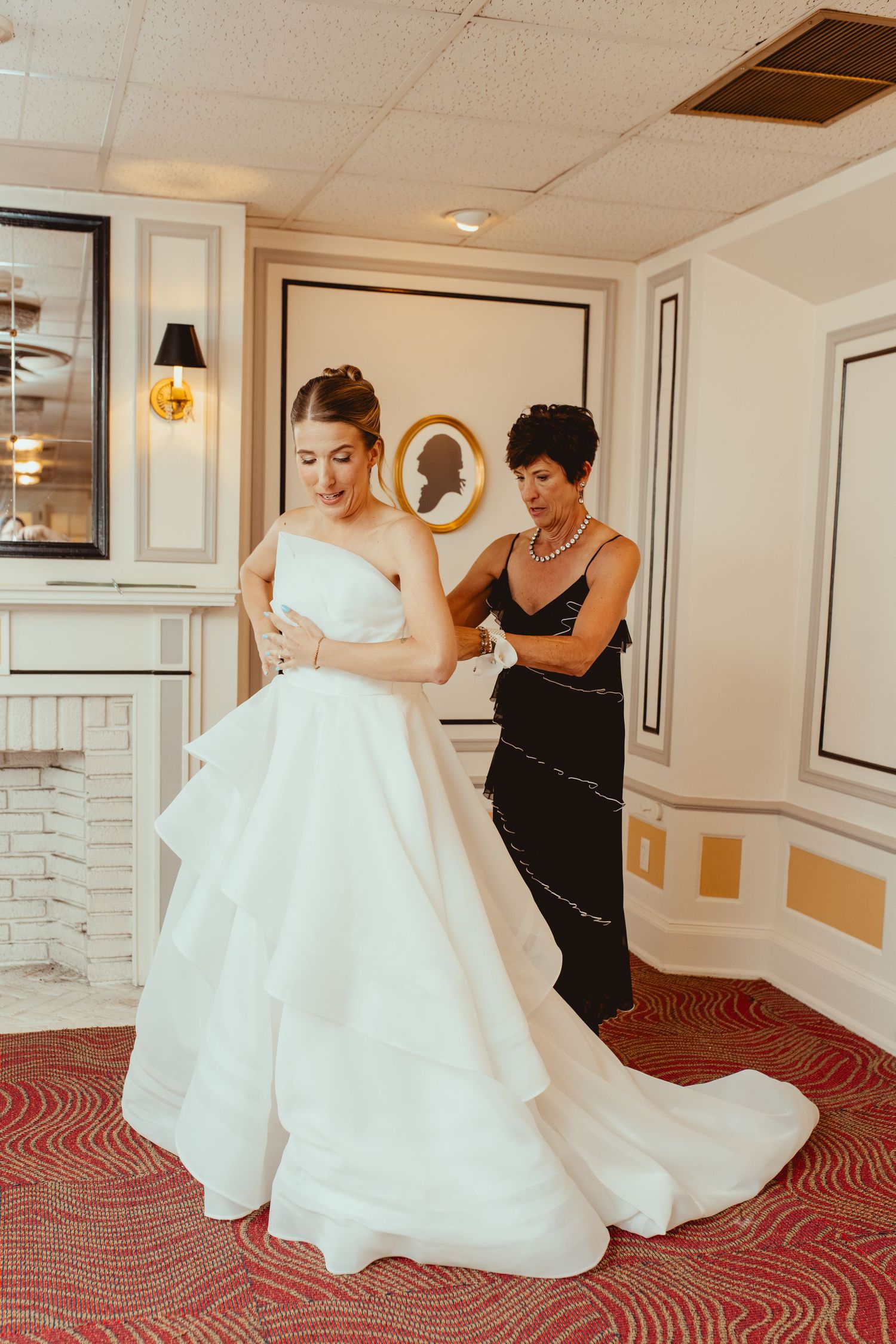 Wedding dress being adjusted on red patterned carpet in elegant hallway.