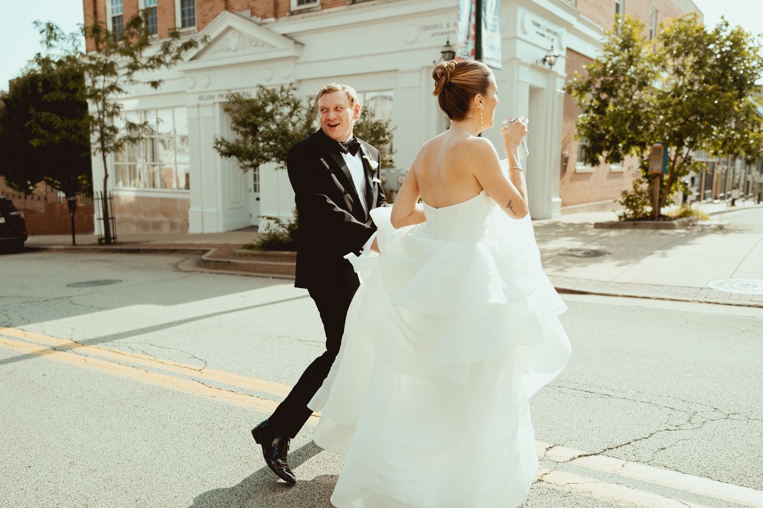 A couple in wedding attire playfully runs down a city street while being photographed.