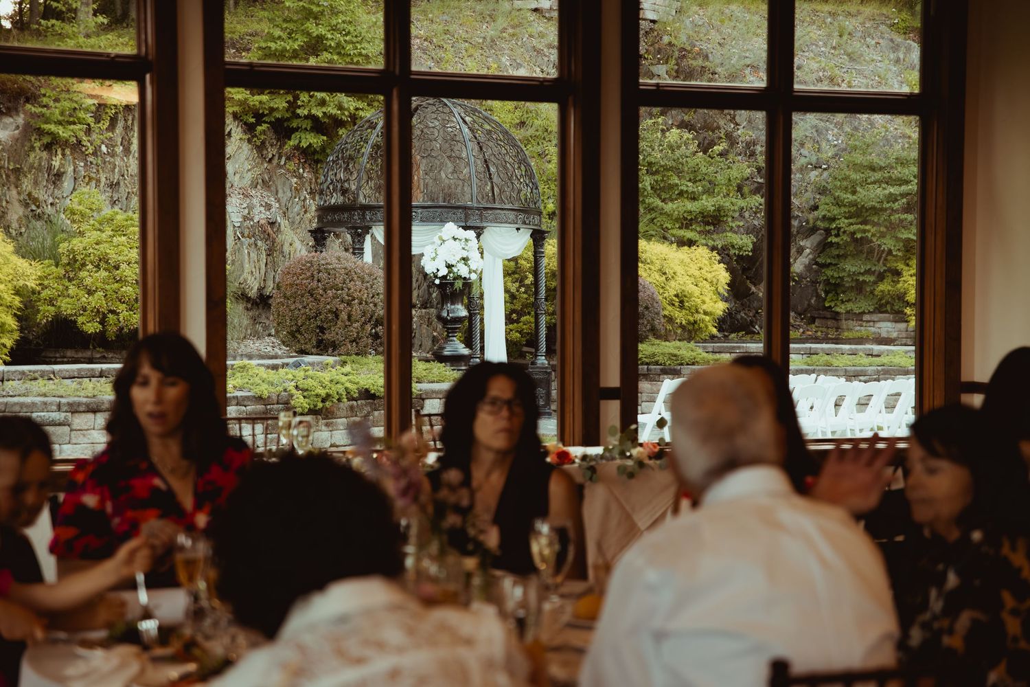 Wedding reception guests dining near large windows overlooking a lush garden gazebo.