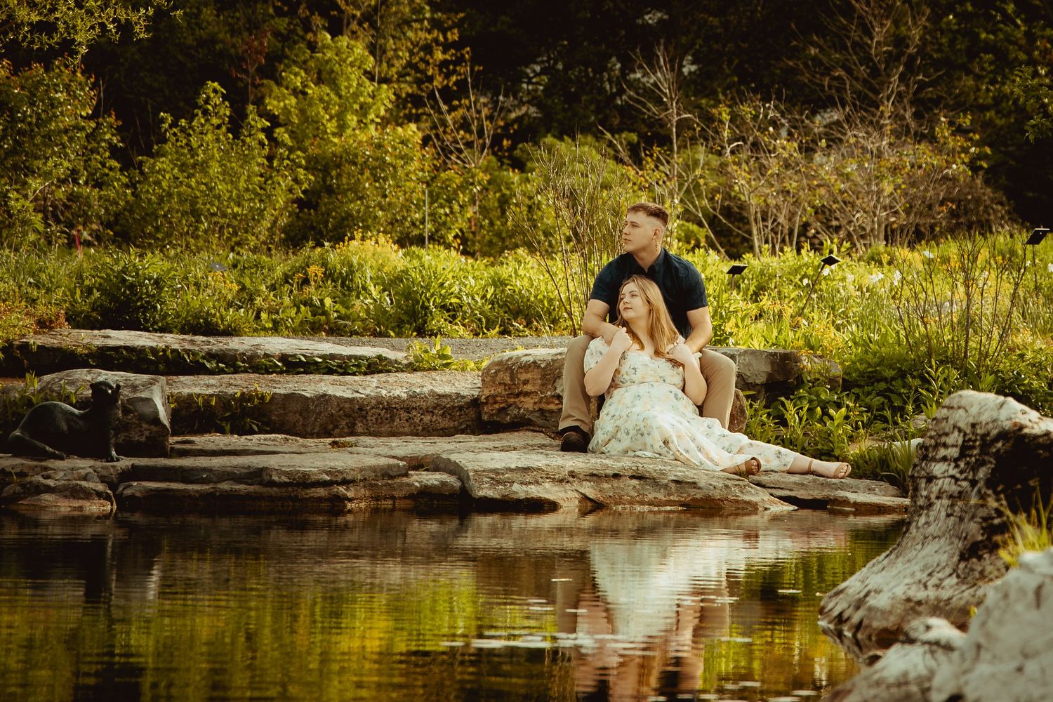 Couple sitting on rocks by a reflective pond surrounded by lush green trees in a romantic outdoor setting.