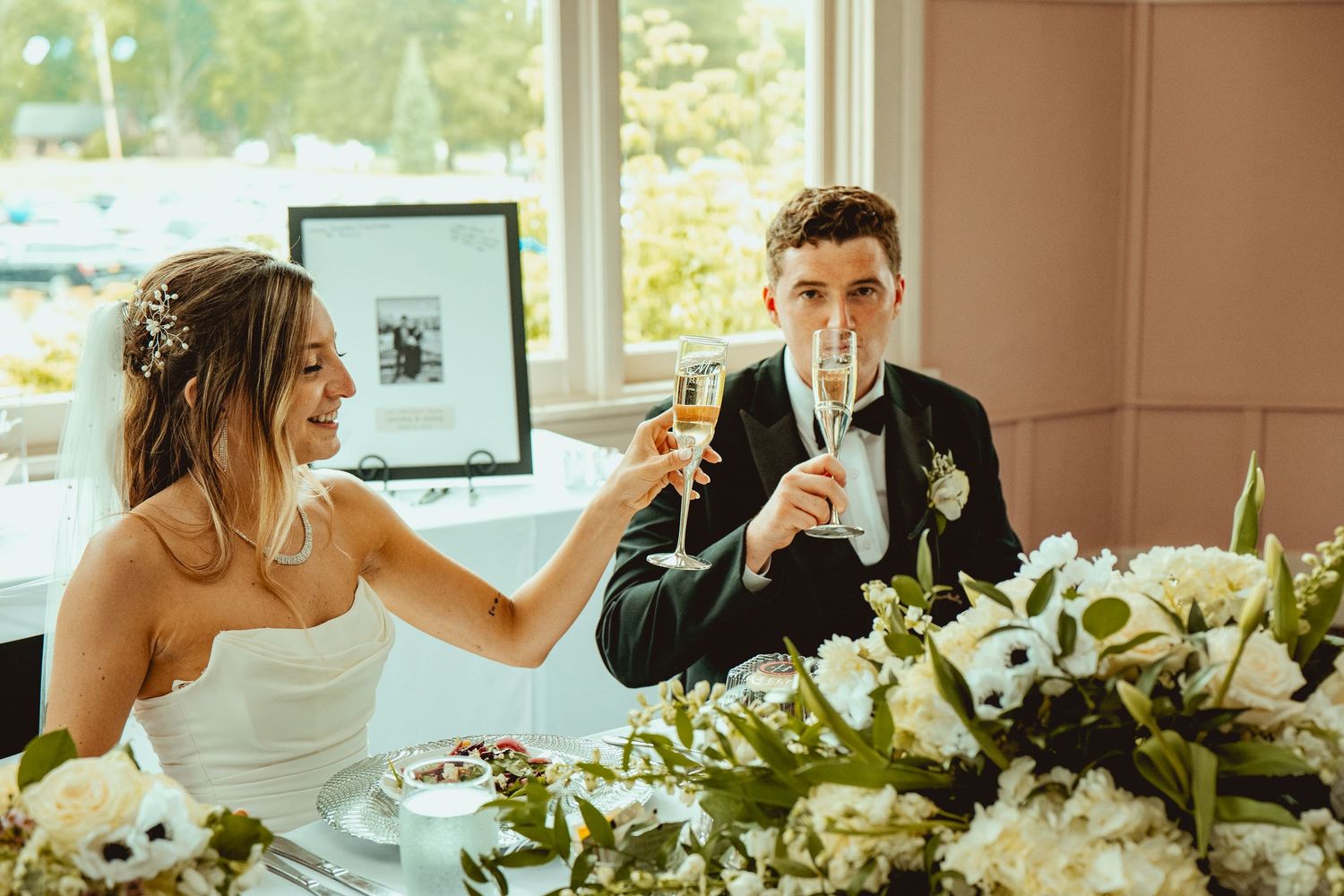 Wedding reception table decorated with white floral arrangements near sunny windows.