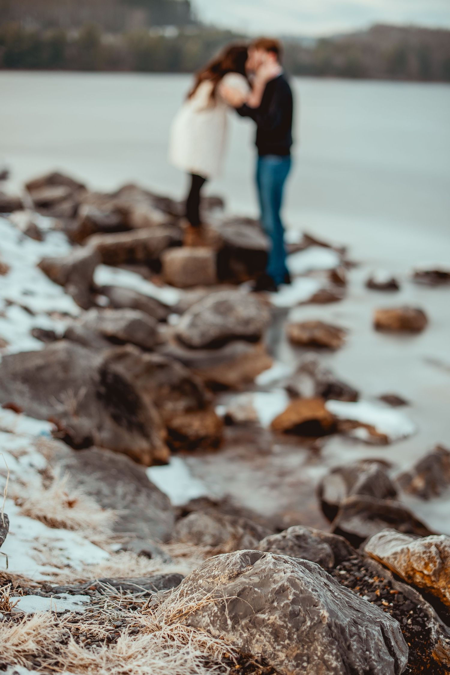 Rocky shoreline with scattered boulders and driftwood along a misty waterfront with figures in the background.
