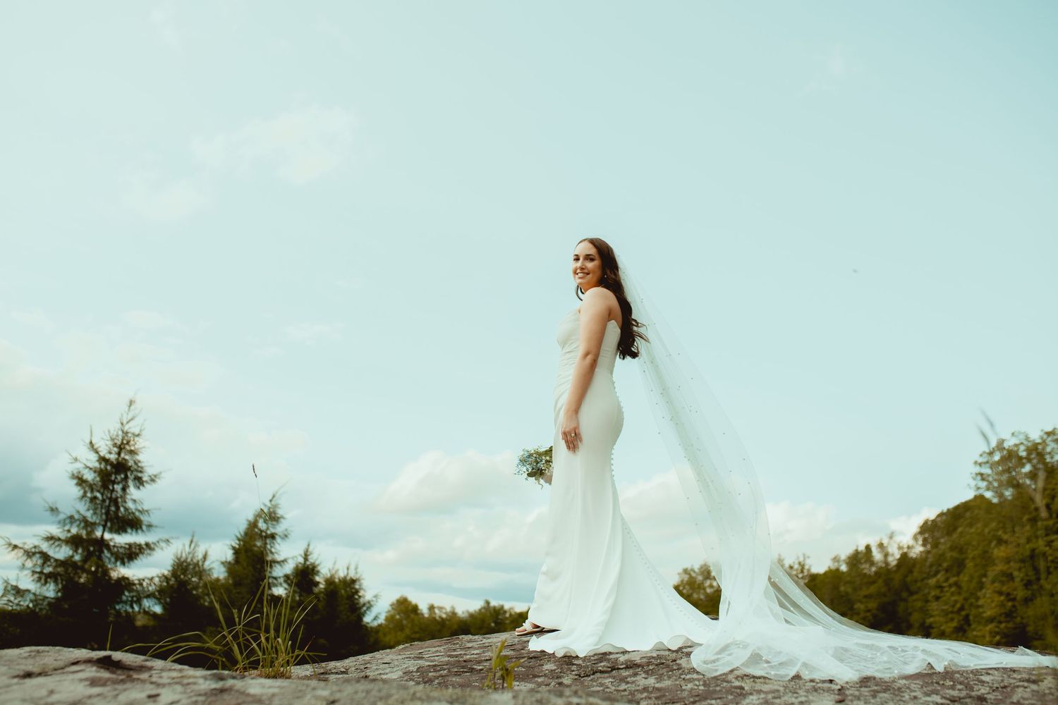 A bride in a flowing white dress and long veil poses dramatically on a rocky outcrop against a blue sky.
