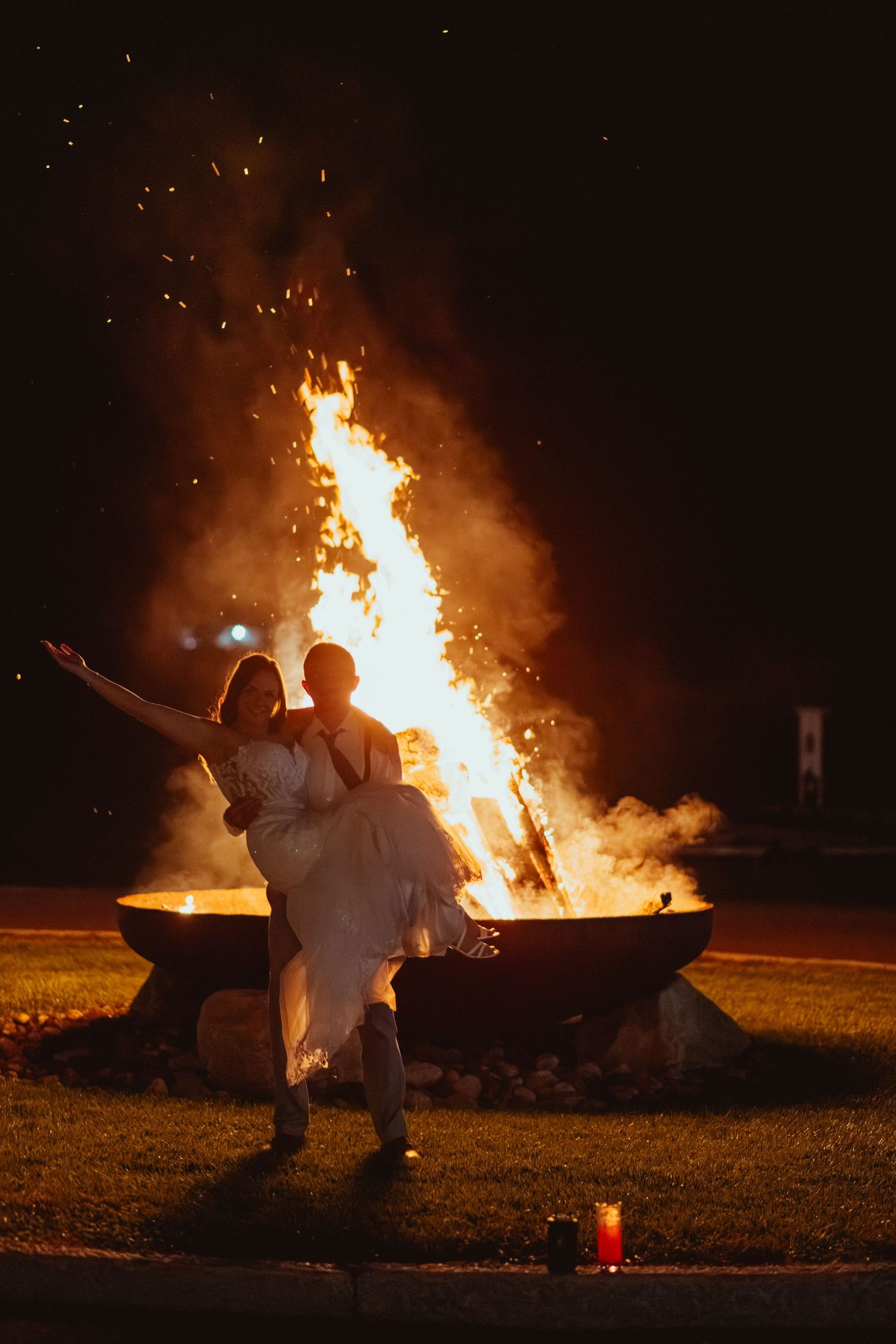 Silhouettes of people sitting near a large bonfire at night with sparks flying in the dark sky.