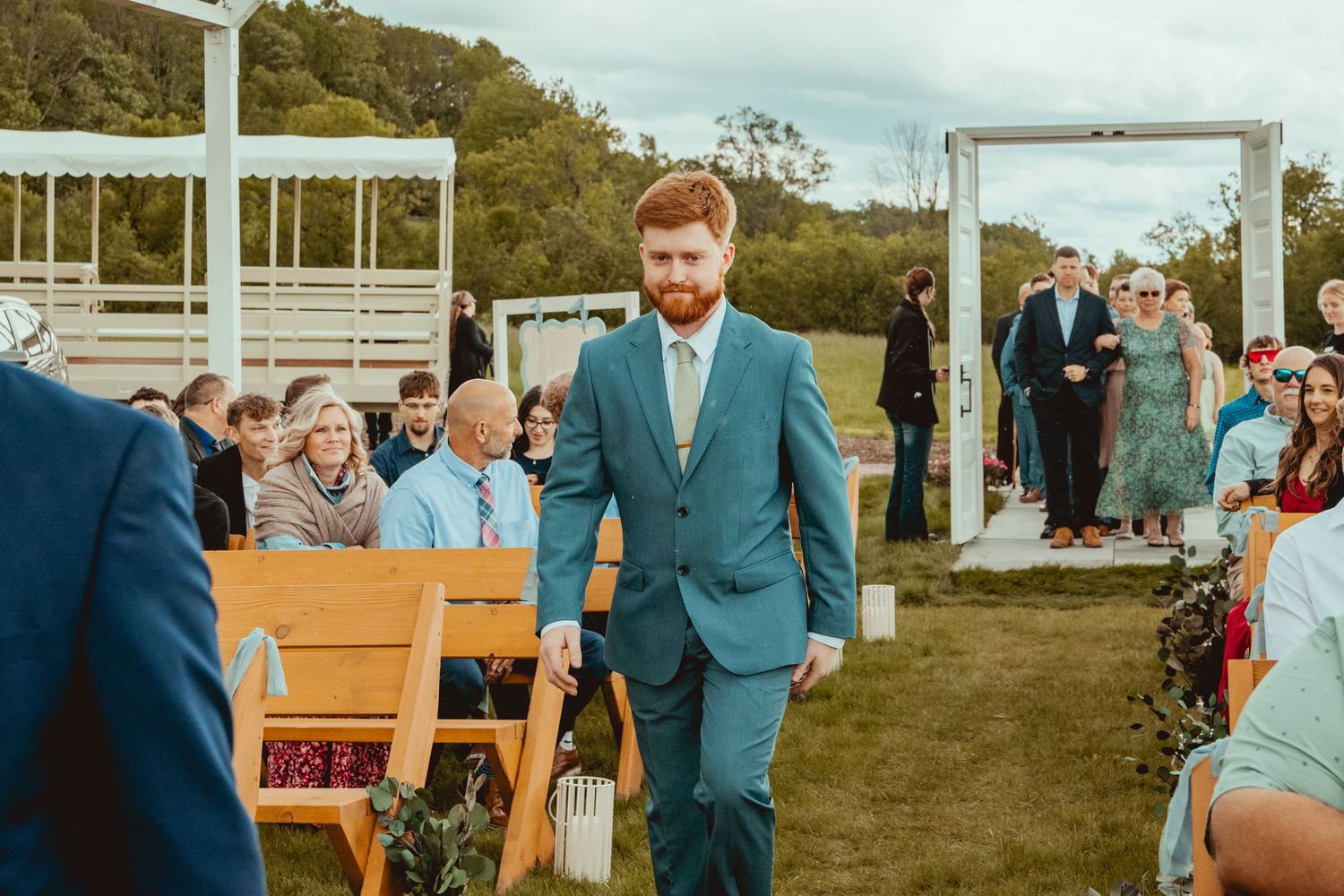 Guests seated on wooden benches at an outdoor wedding with a white ceremonial arch.