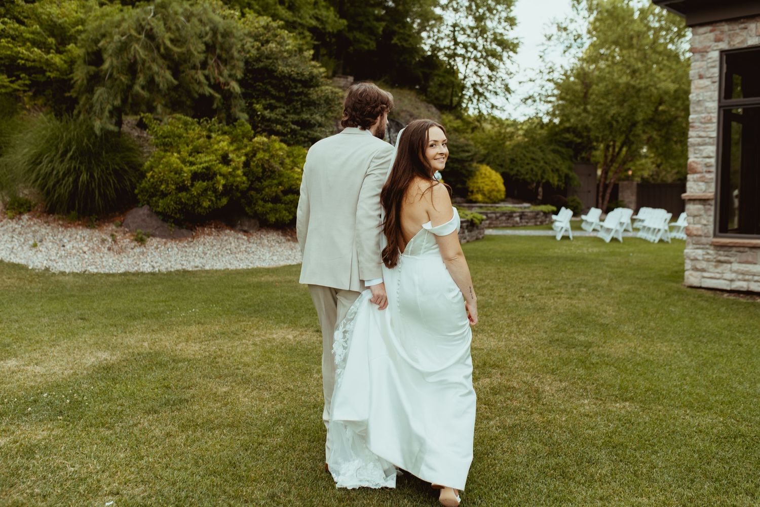 Bride in light blue gown walks hand-in-hand with groom in beige suit across lush green lawn.