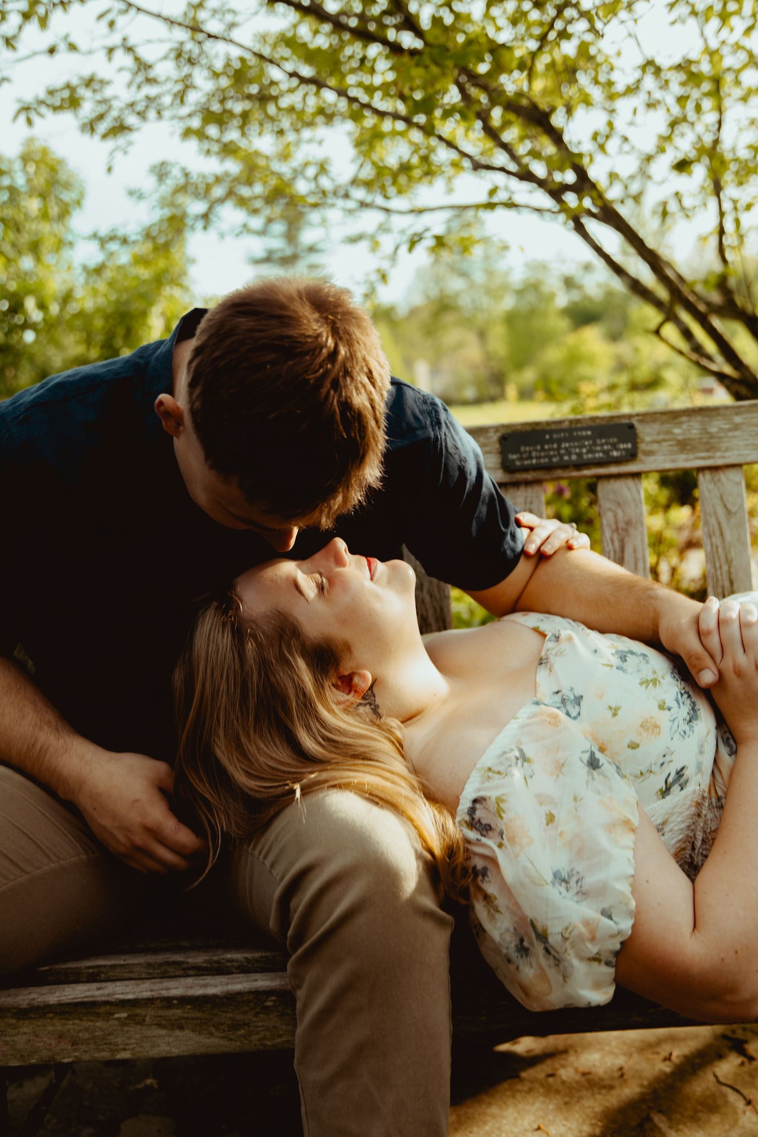 A couple shares a romantic moment on a wooden bench outdoors, with the woman reclining as the man leans in to kiss her.