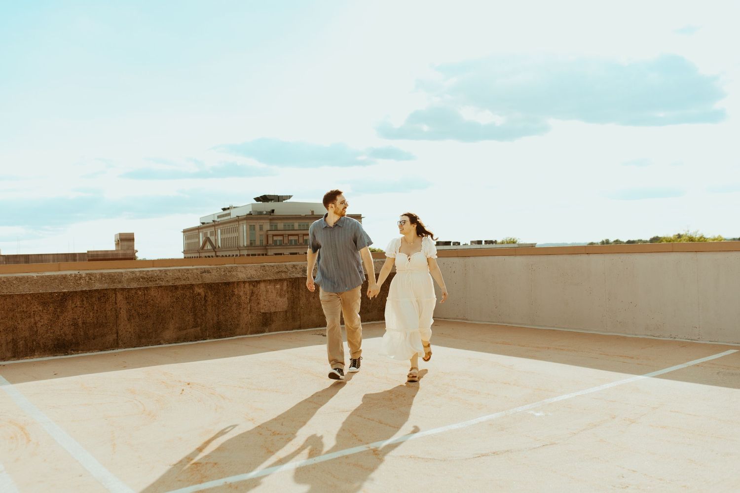 Couple holding hands walking on rooftop under blue sky, wearing casual white and gray outfits on a sunny day.