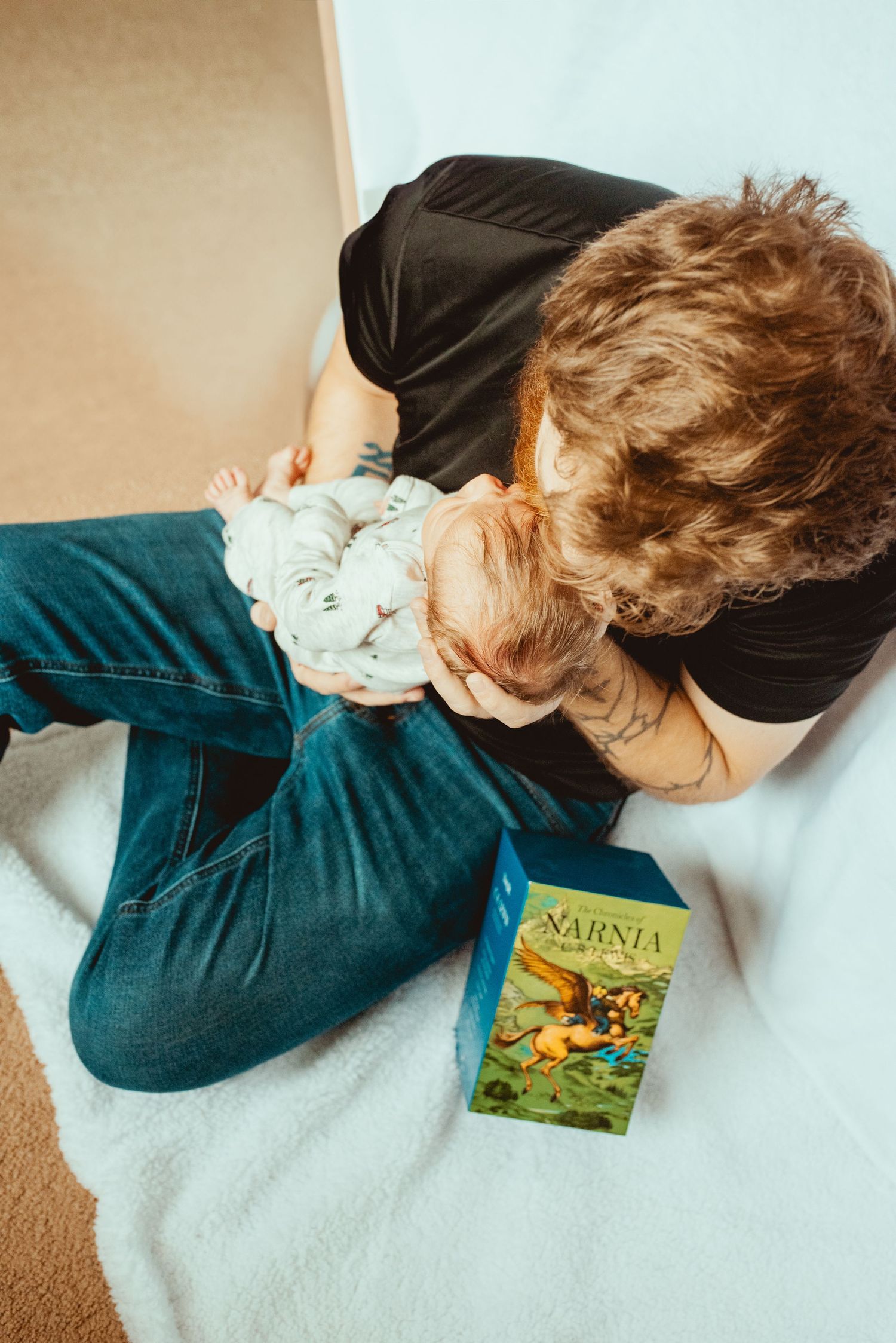 A person in dark clothing lies curled up on white bedding next to a Narnia book, creating a cozy reading scene.
