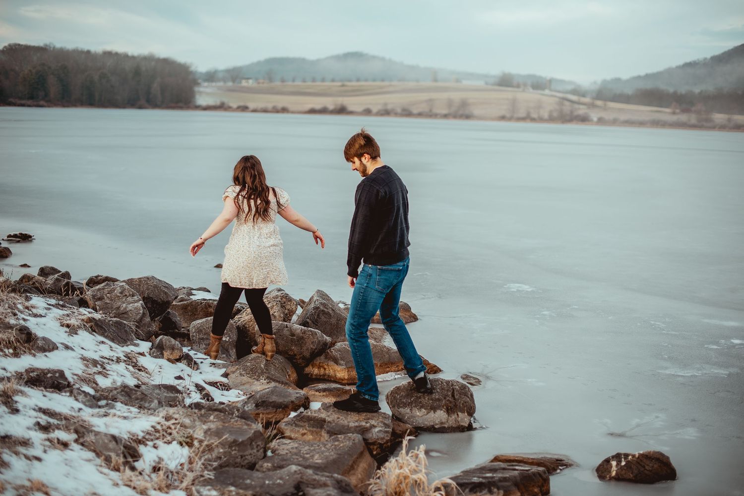 Couple walks along rocky shoreline with mountains and cloudy skies in the background.
