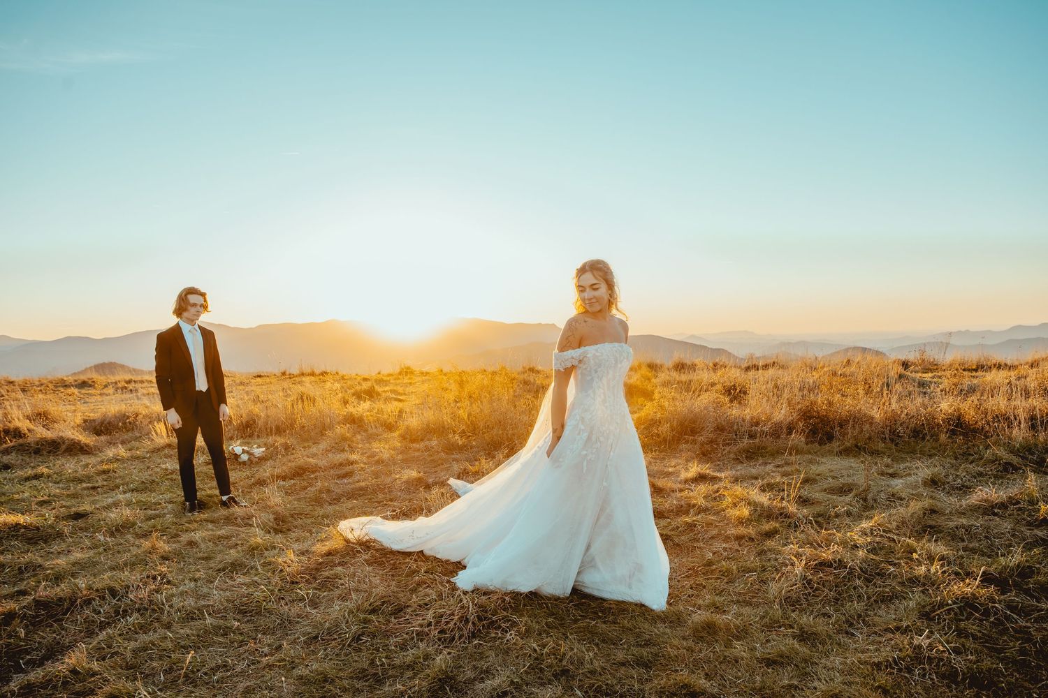 A couple in wedding attire stands in a golden field at sunset with mountains in the background.
