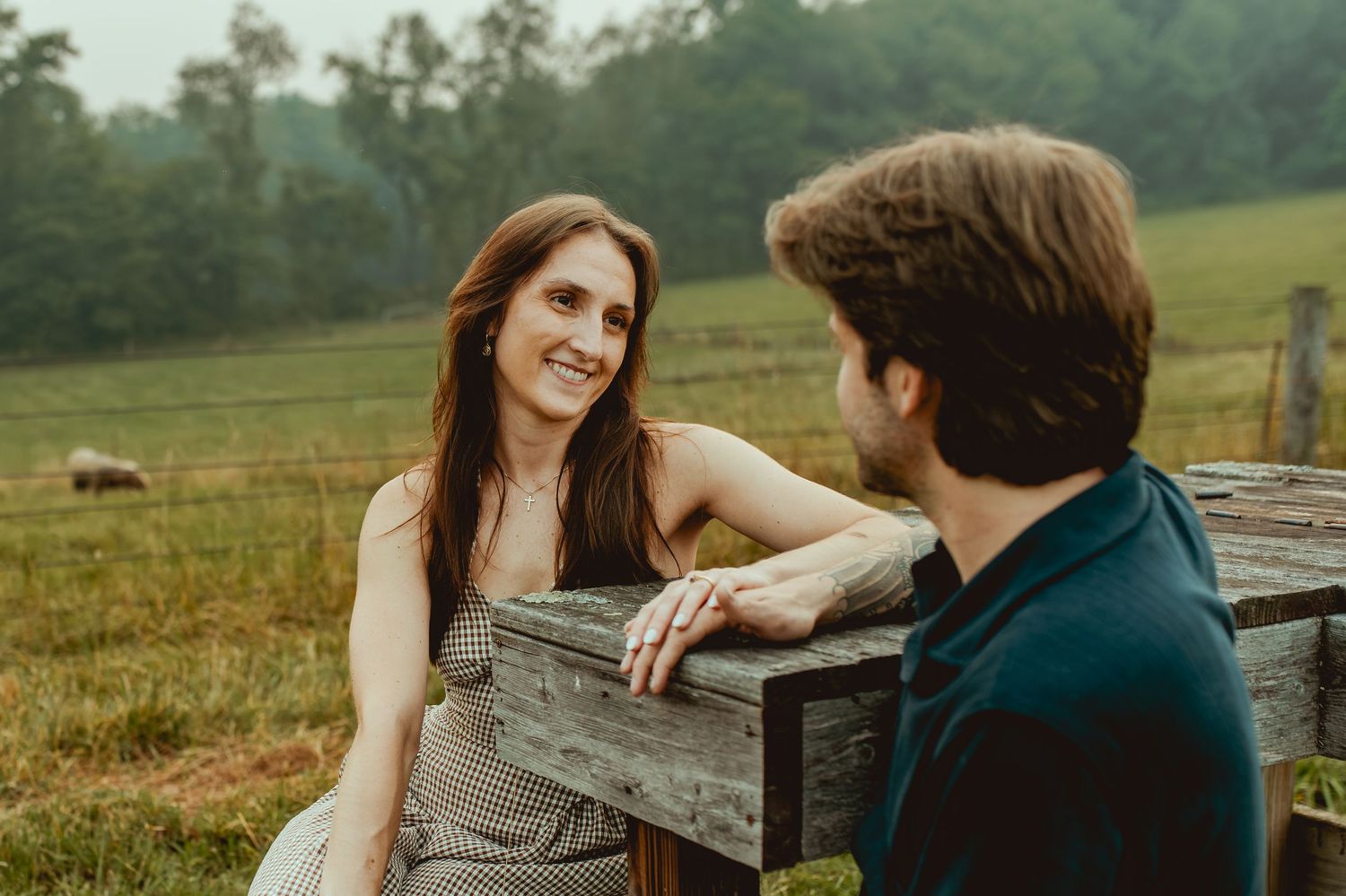 Couple sharing a romantic moment by a rustic wooden fence in a lush green countryside field.