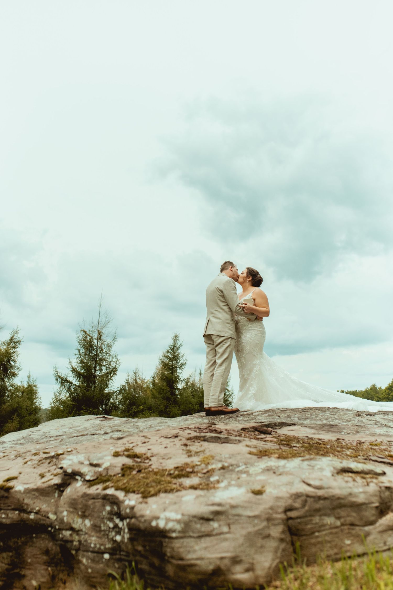 Bride and groom kissing on a rocky cliff surrounded by pine trees under a cloudy sky on their wedding day.