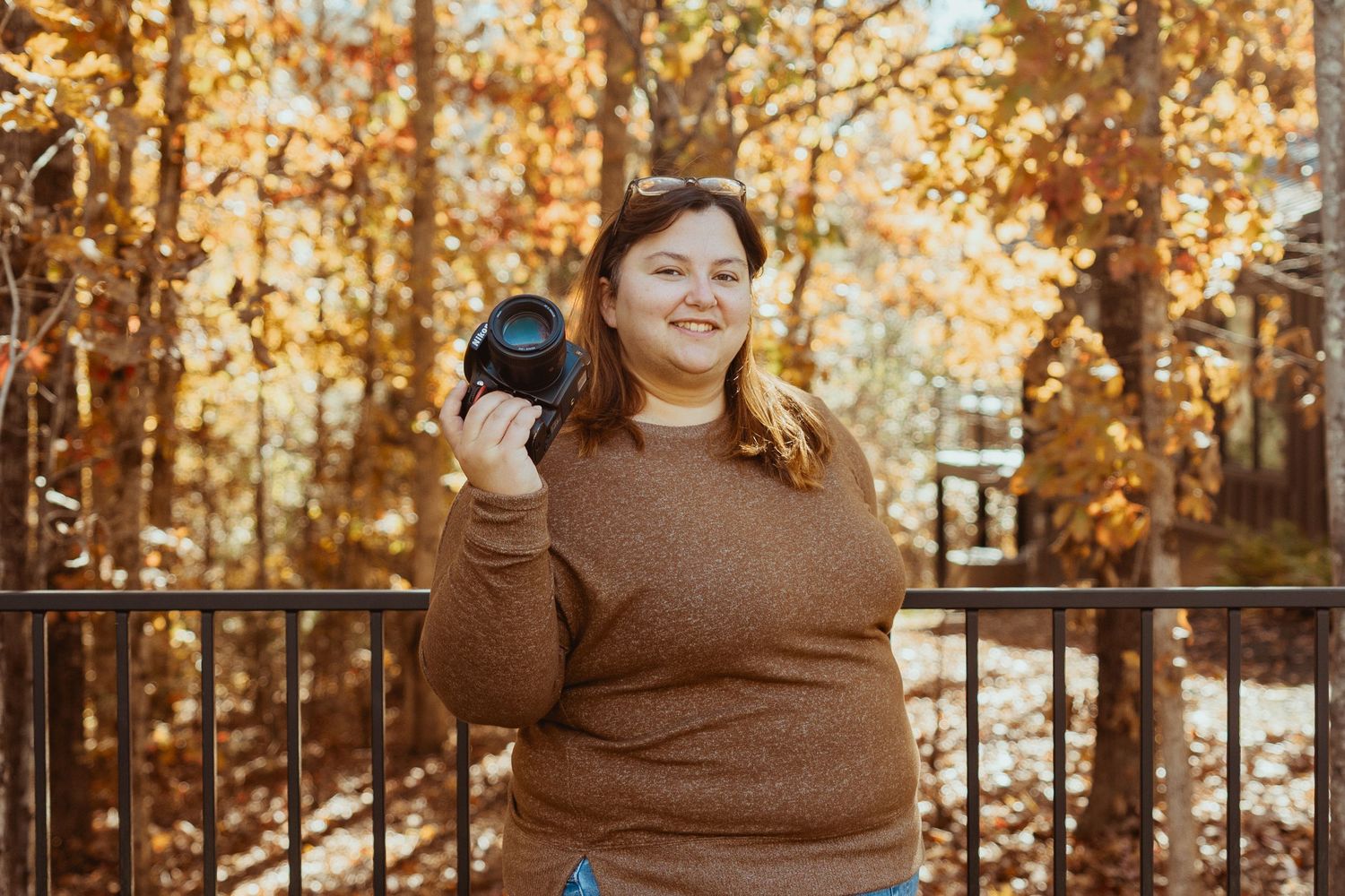 Young woman holding a camera outdoors in autumn, surrounded by golden fall foliage and a wooden fence.