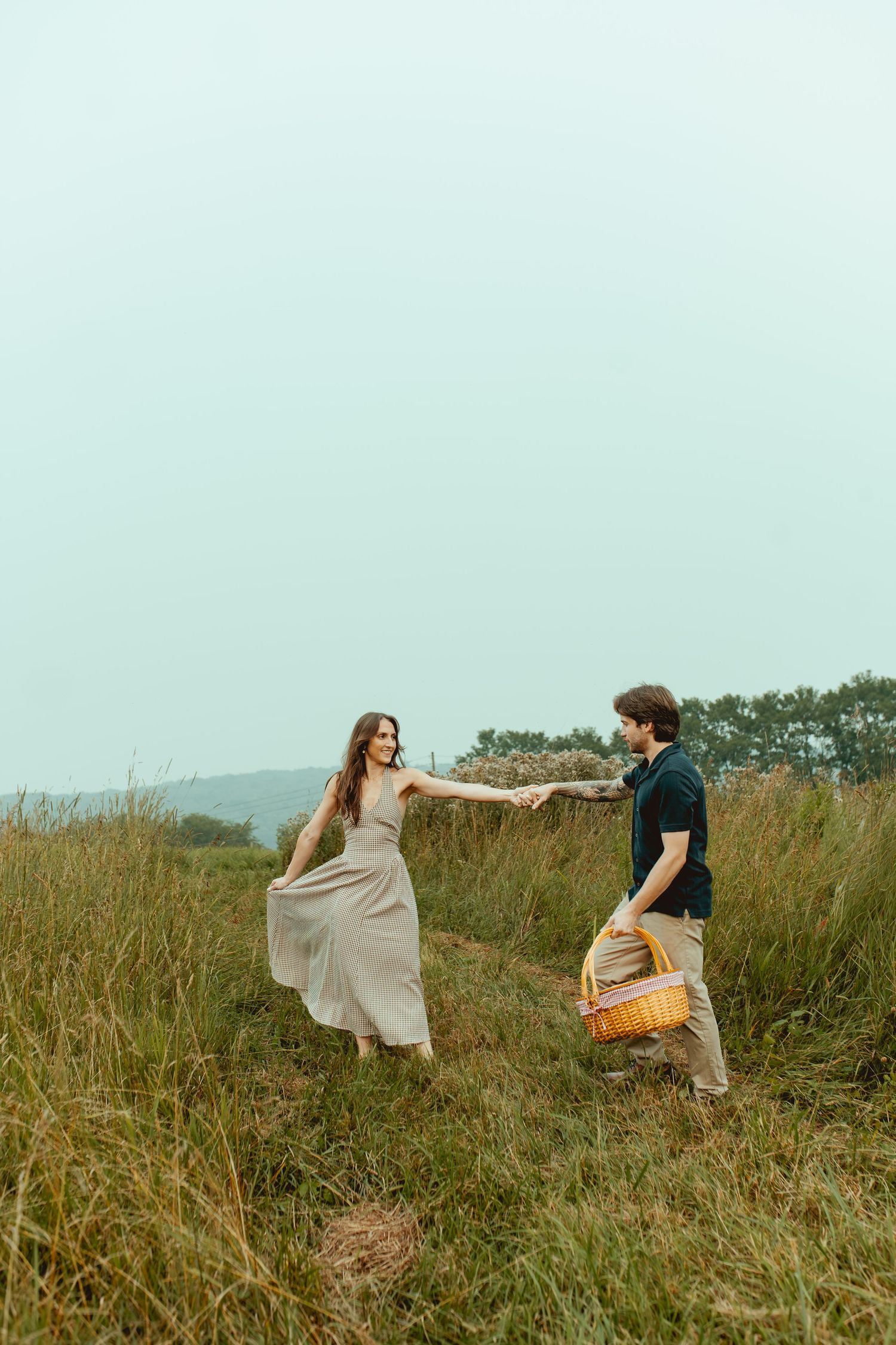Couple holding hands in a grassy field, woman in flowy dress, man carrying a picnic basket on a cloudy day.