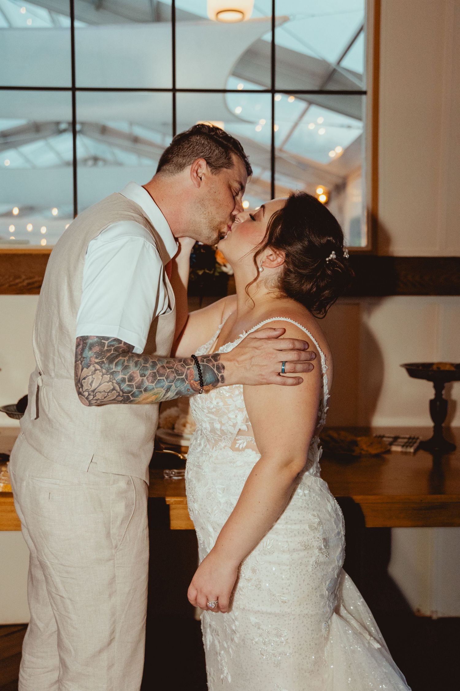 Tattooed groom kisses bride in white lace dress at rustic indoor wedding reception.