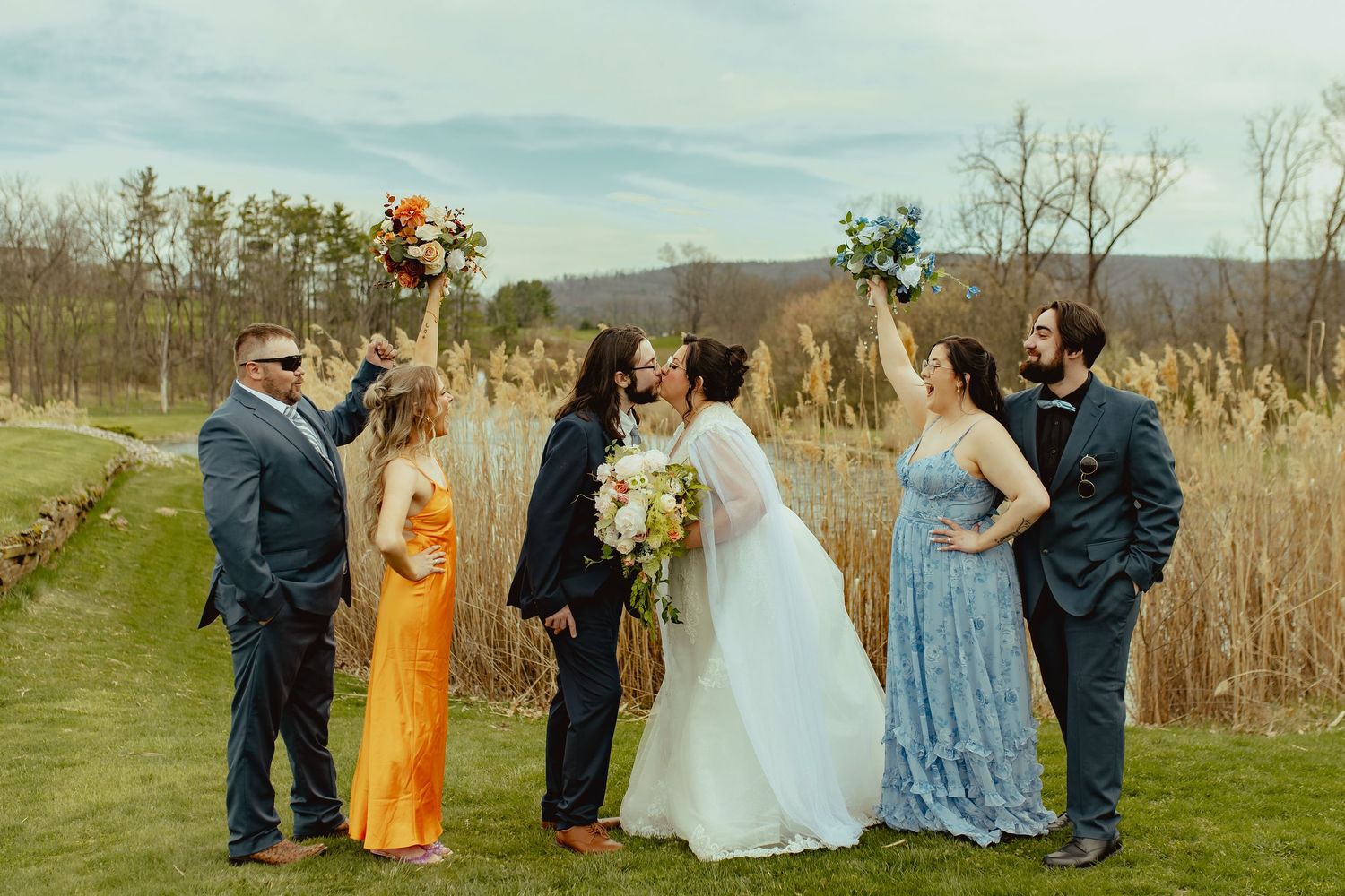 Wedding party celebrates outdoors as bride and groom kiss, surrounded by bridesmaids and groomsmen raising bouquets.