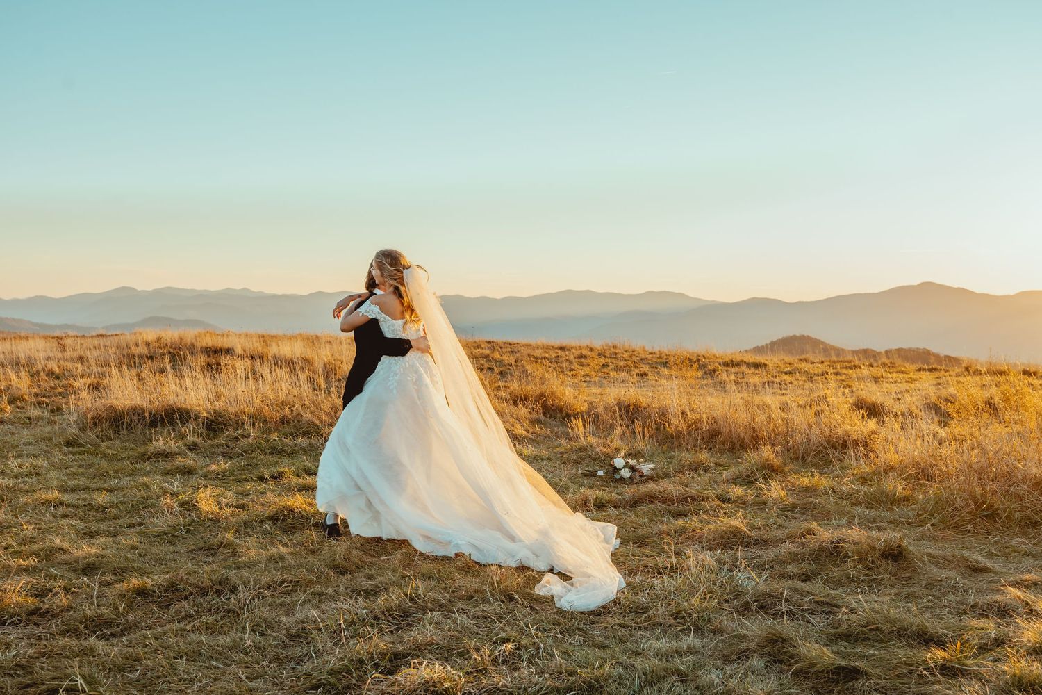 Bride in flowing white dress twirling in mountain field at sunset.