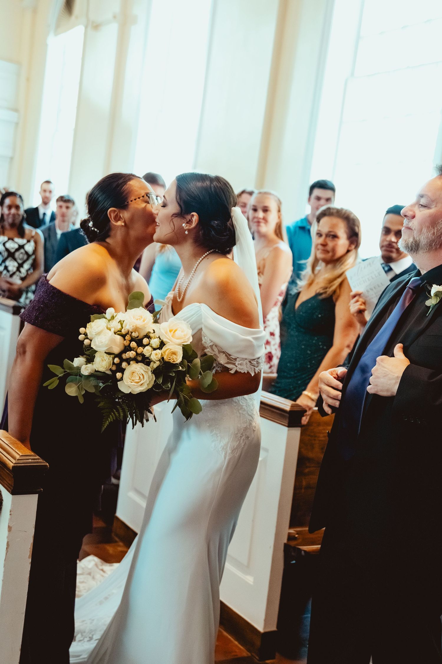 Wedding ceremony guests embrace and share joyful moments in a bright church setting with white pillars.