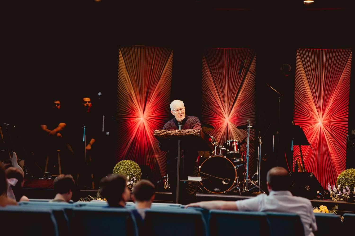 A speaker stands at a podium on stage with dramatic red lighting and an audience visible in the foreground.
