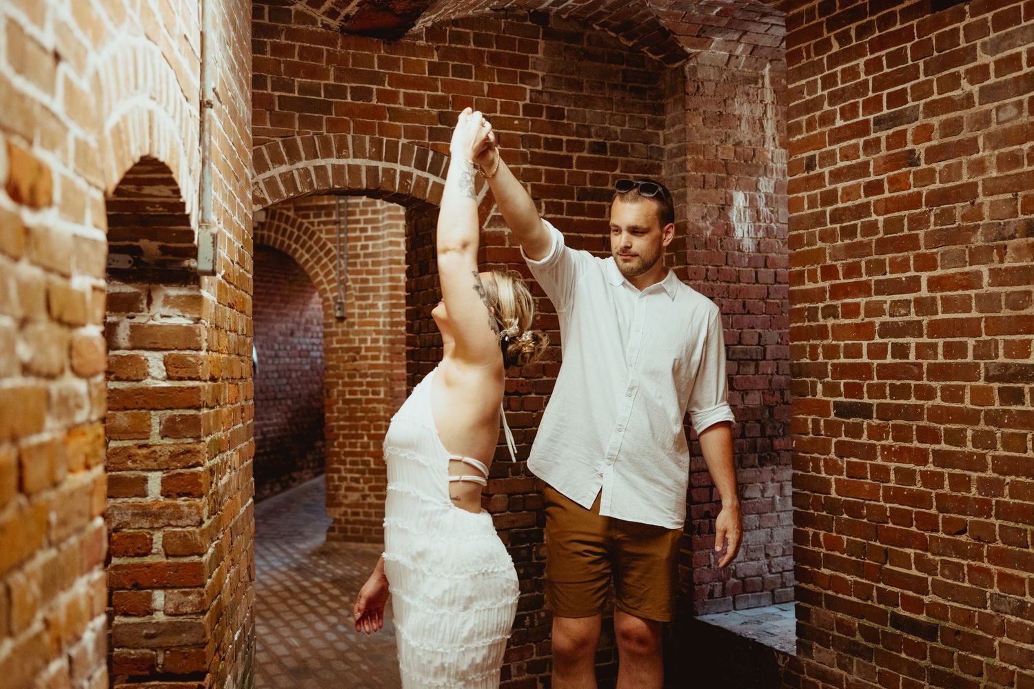 Couple dancing in rustic brick archway, bride in lace dress, groom in white shirt and tan shorts.