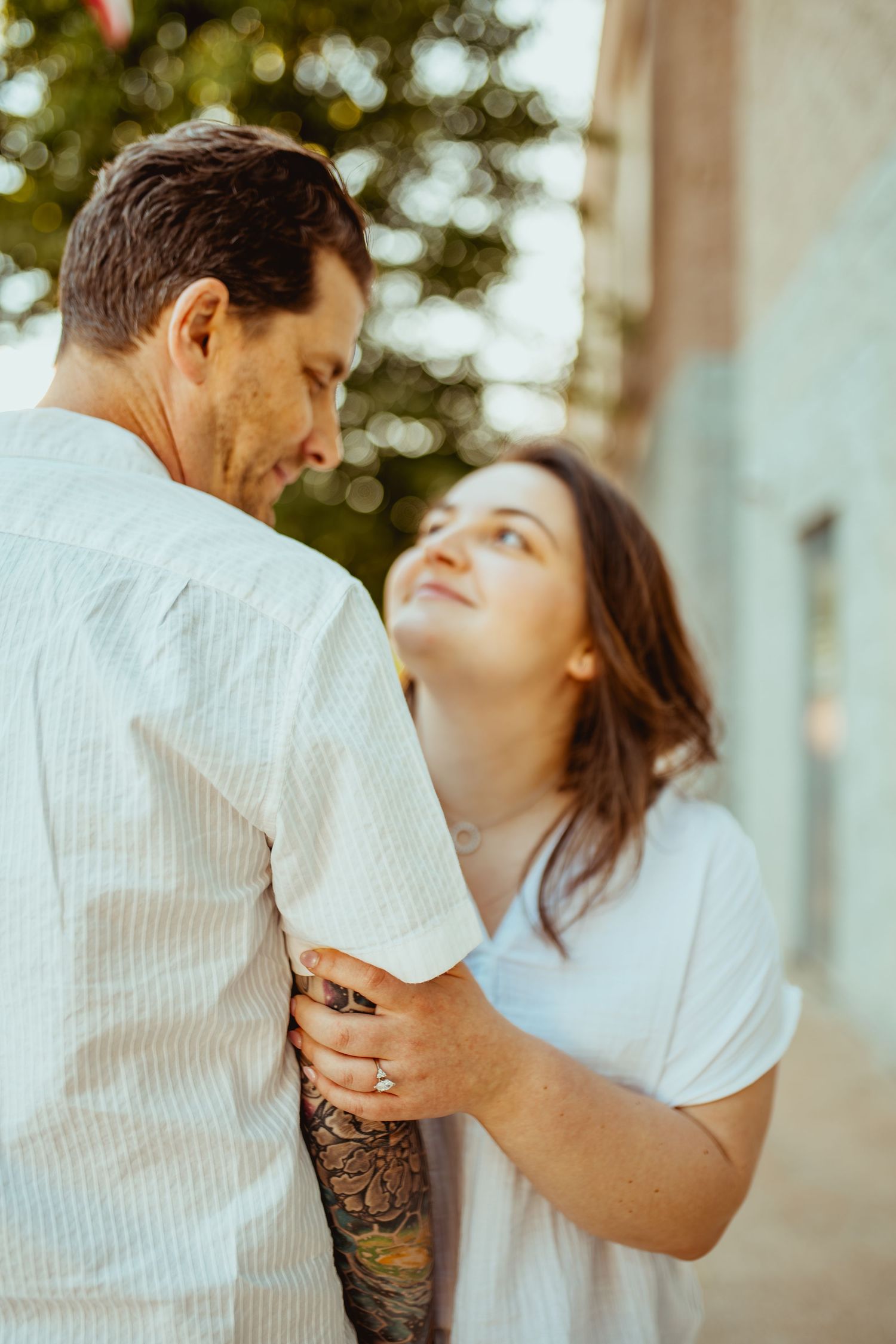 Woman gazes lovingly up at tattooed man while holding his arm during outdoor couples engagement session.