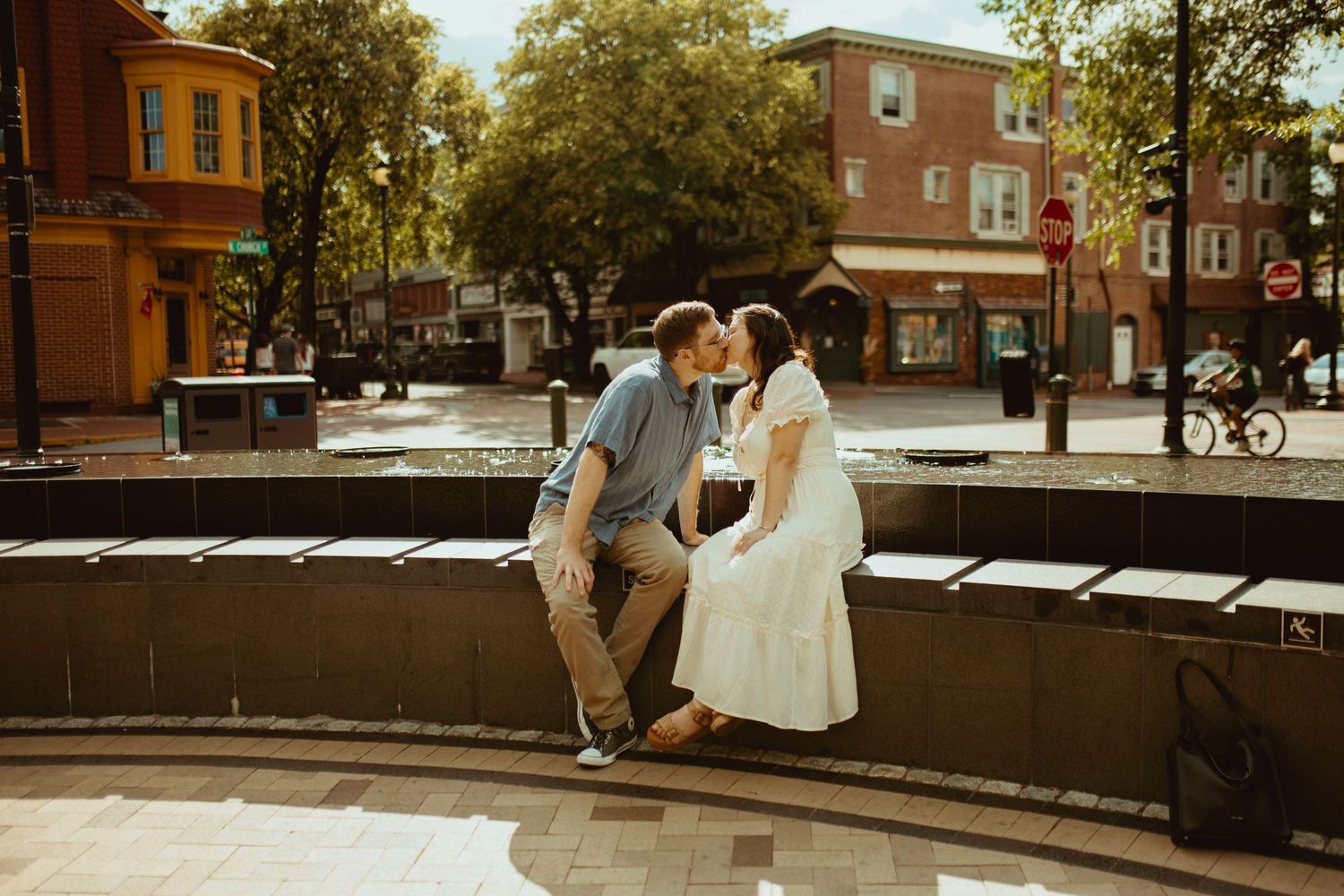 Couple sharing a romantic kiss while sitting on a fountain ledge in a charming urban plaza surrounded by brick buildings.