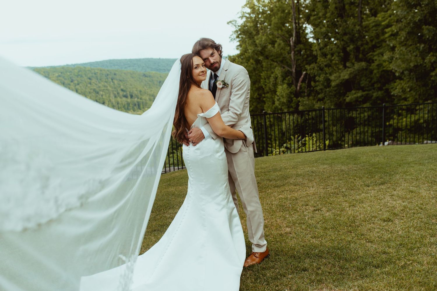 Bride in fitted white gown with flowing veil poses with groom against lush green hillside.
