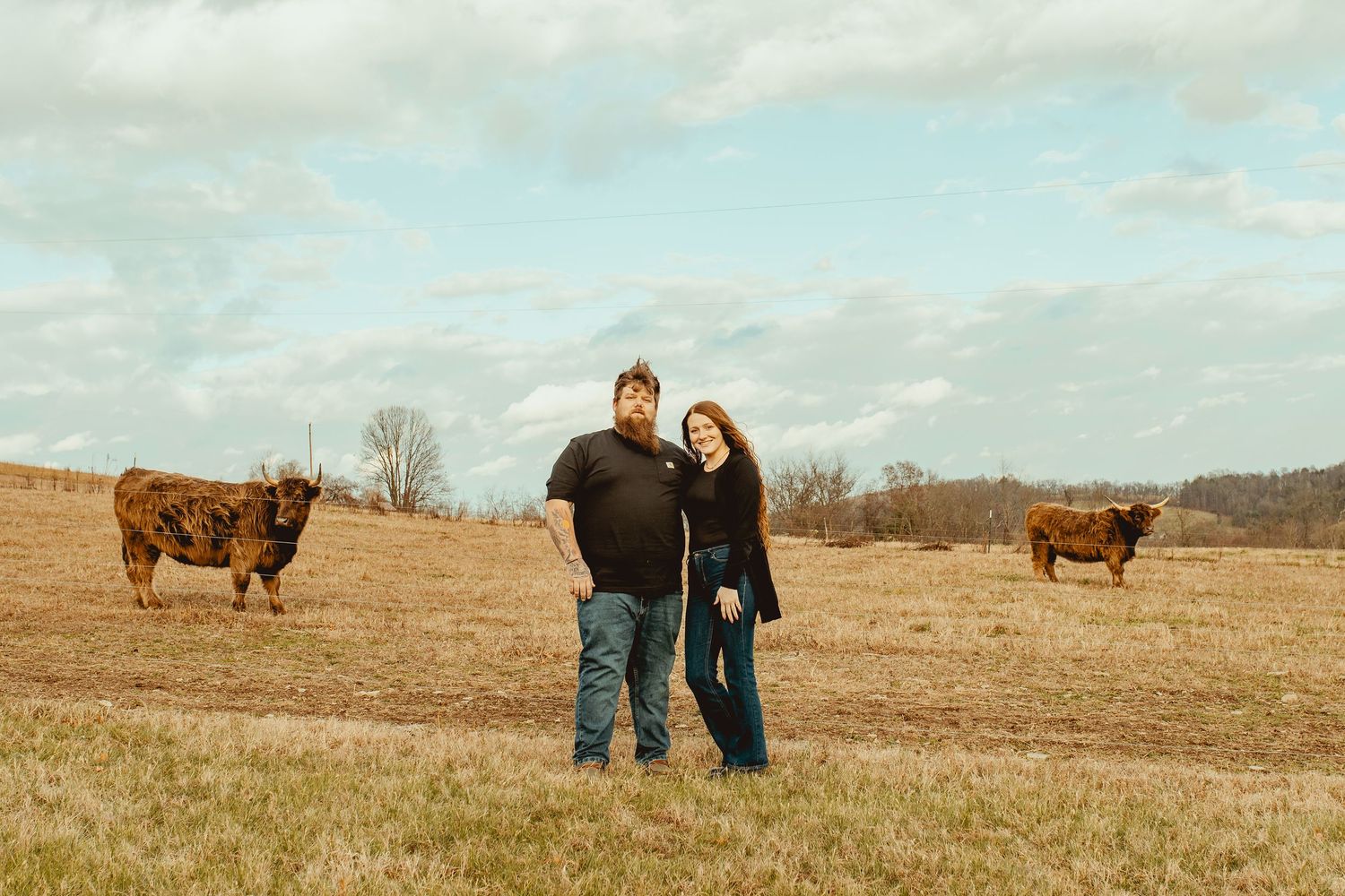 A couple poses together in a field with Highland cattle grazing in the background on a cloudy autumn day.