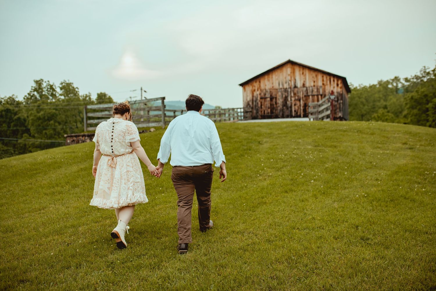 A couple walks hand in hand towards a rustic barn on a grassy hillside.
