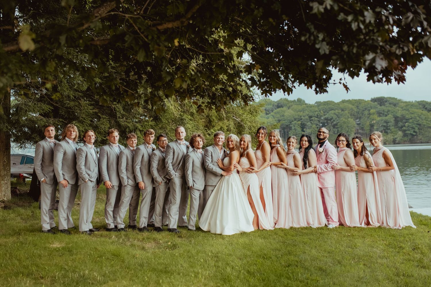 Wedding party poses under tree by lake, groomsmen in gray suits and bridesmaids in blush gowns.