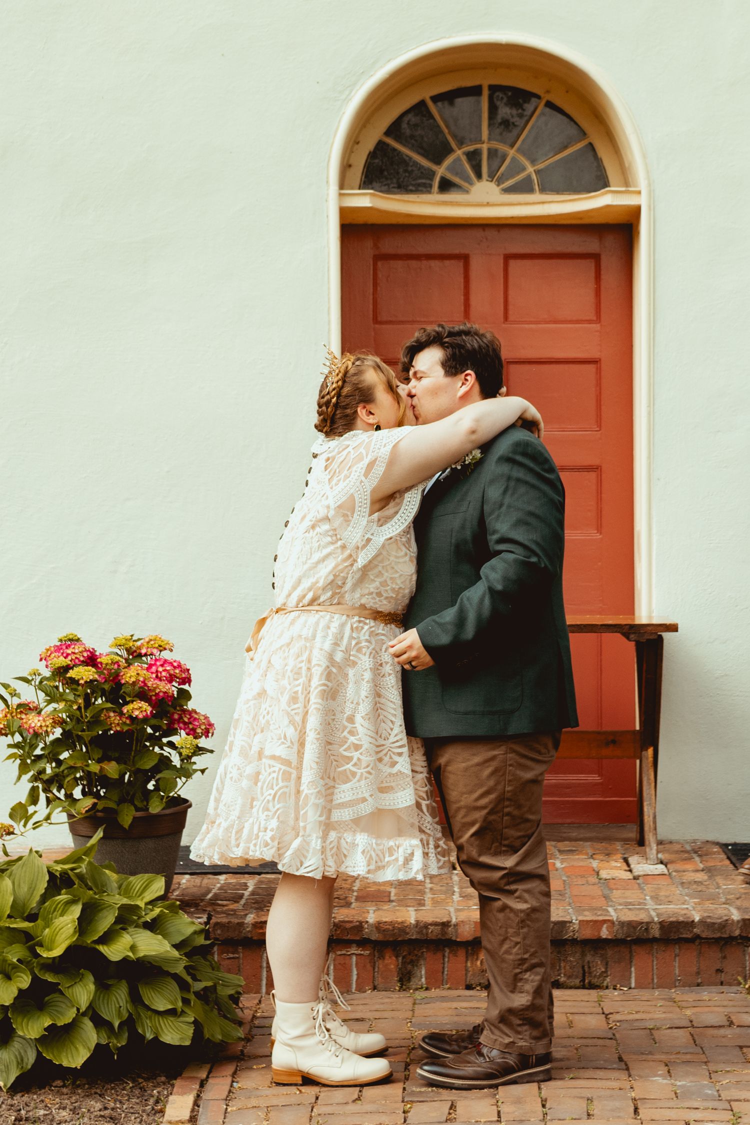 A romantic couple shares a tender kiss on a brick patio in front of a red door and arched window.