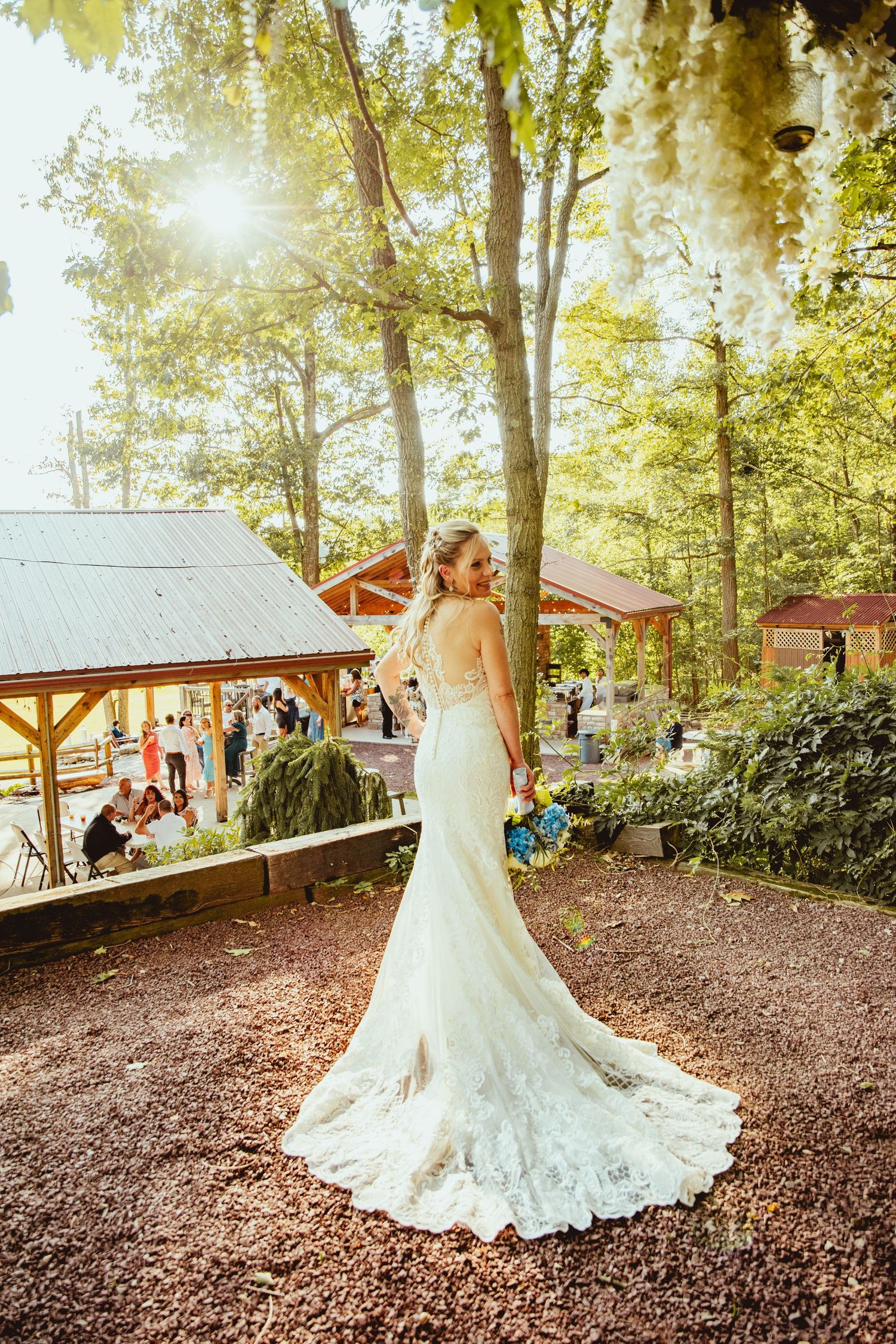 Bride in lace wedding dress poses in a sunlit forest setting with rustic buildings in background.